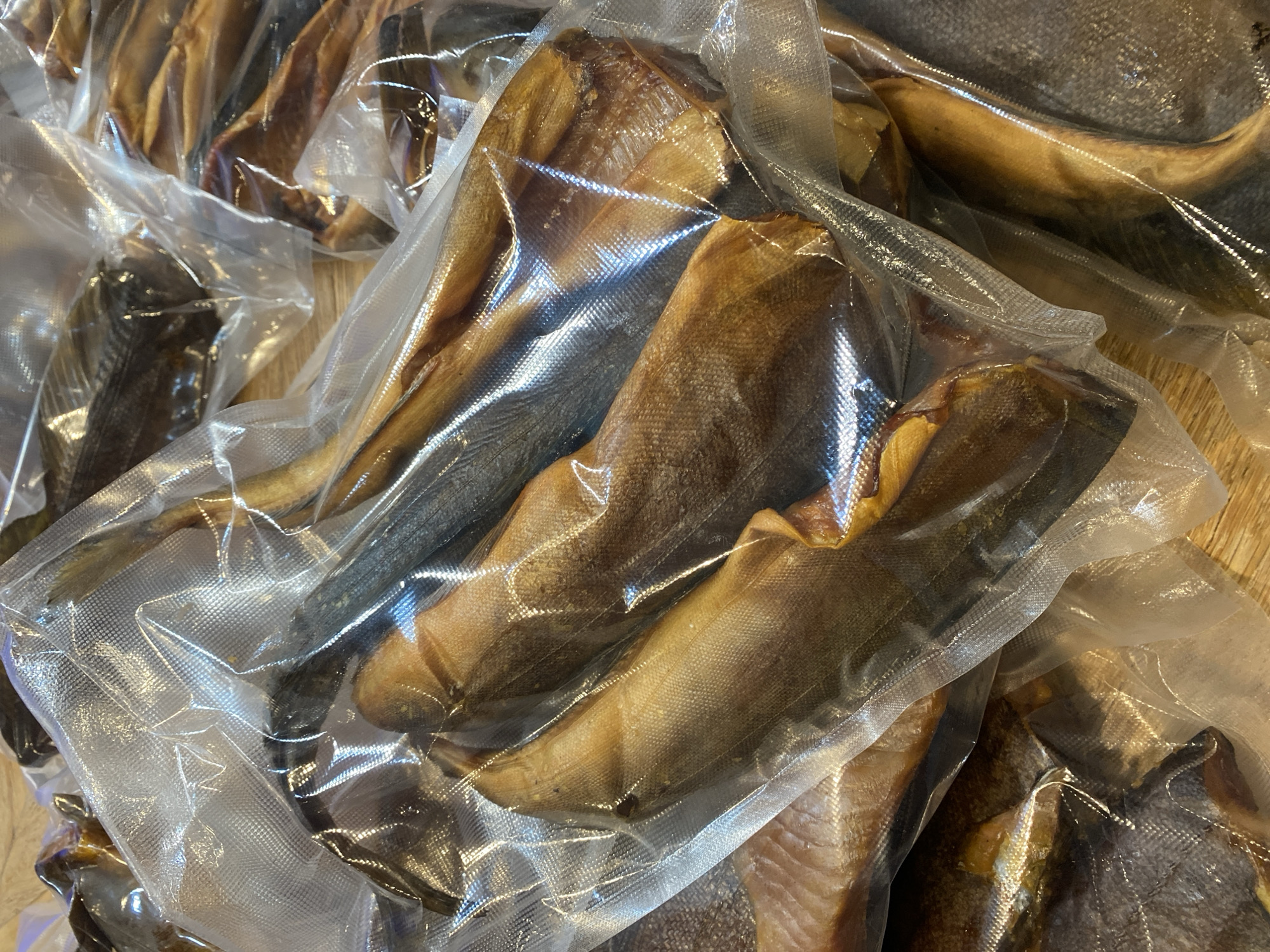 The image shows several vacuum-sealed packages of smoked fish, likely herring or a similar small, oily fish, arranged on a wooden surface. The fish are golden brown and appear to be whole. The packaging is clear plastic with a textured sealing strip. The lighting suggests an indoor setting, possibly a market or a home kitchen in Tret'ya Pad', Russia, given the context. There are no people visible in the image. The focus is on the preserved fish products.