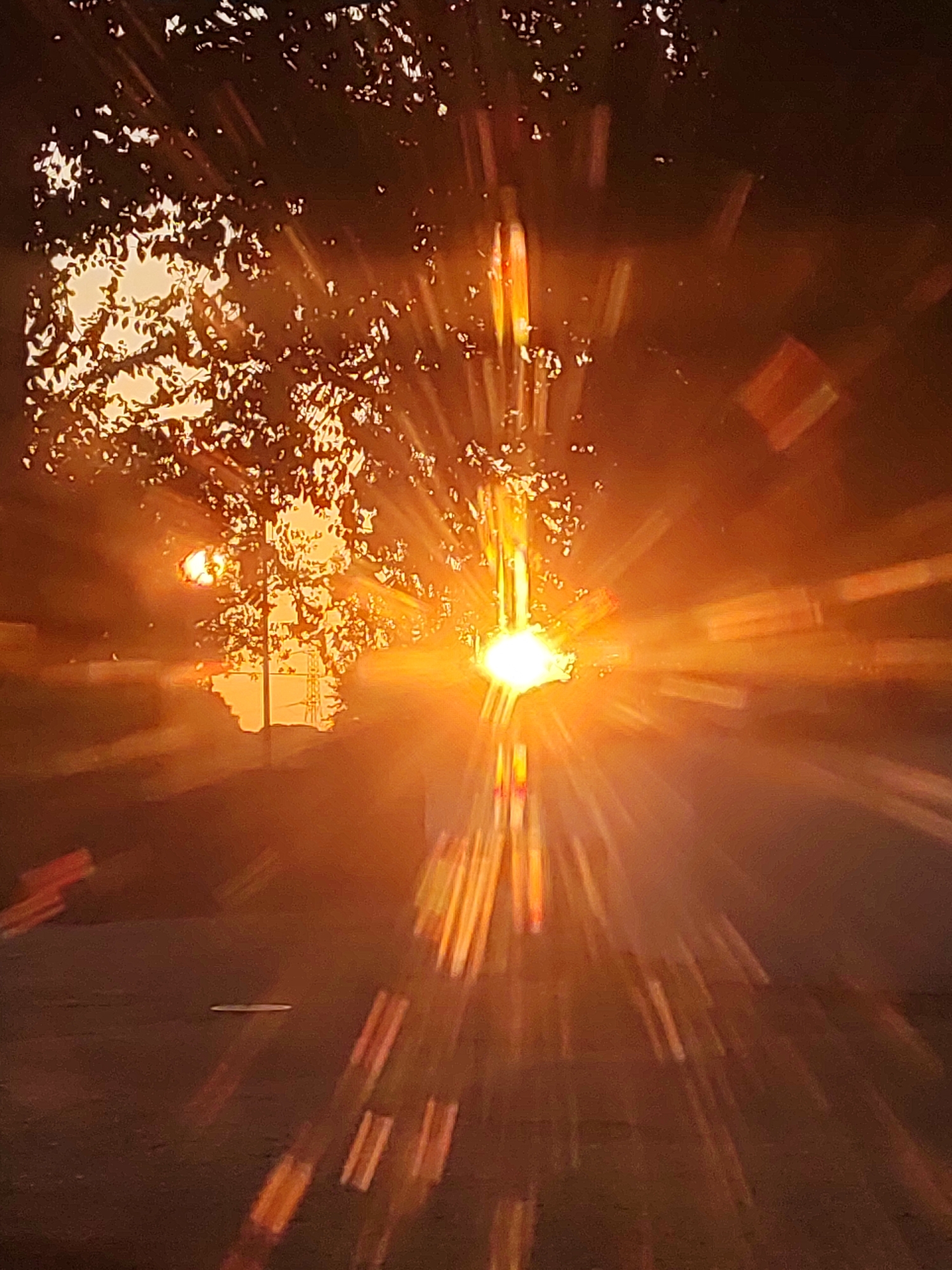 The image depicts a sun flare effect, likely captured through a window or camera lens, with prominent streaks of light emanating from a bright central source. The scene appears to be outdoors, possibly in Traspinedo, Spain, given the context of the query. In the upper left portion of the image, tree branches with leaves are silhouetted against the bright sky, creating a dappled effect where sunlight filters through. A faint structure resembling a utility pole or tower is visible in the background, partially obscured by the foliage and lens flare. The ground in the foreground is dark and appears to be asphalt or a similar surface, with distorted reflections of light streaks. The overall atmosphere is warm and intense due to the strong sunlight and the lens flare effect.