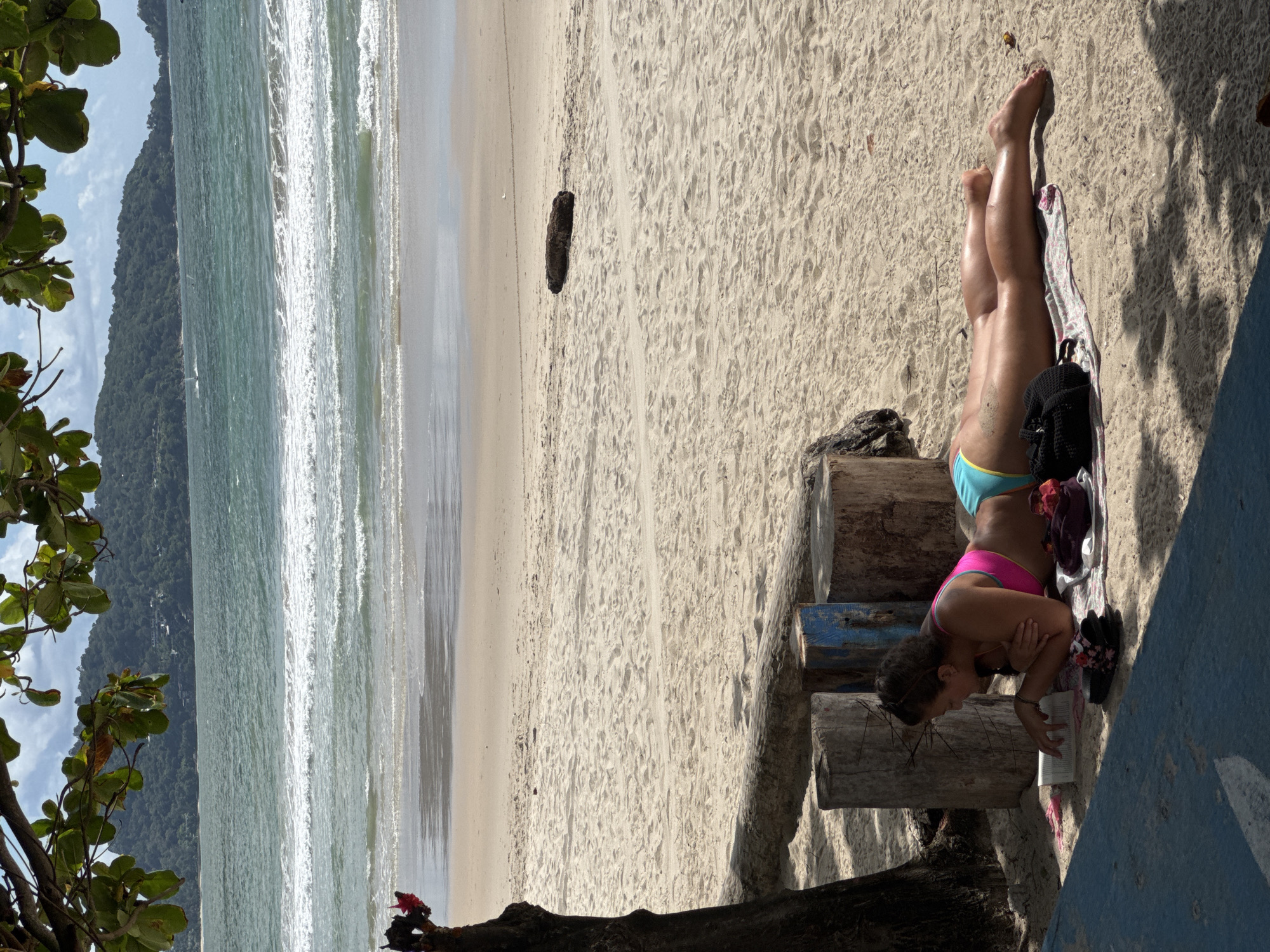 A woman is lying on a towel on a sandy beach, reading a book. She is wearing a pink bikini top and blue bikini bottoms. Her legs are extended in front of her, and she is tanned and appears to be sunbathing. To her left, there is a tree trunk and a pile of logs. In the background, the ocean stretches out to the horizon, with gentle waves breaking on the shore. A green, tree-covered hill rises in the distance, under a cloudy sky. The scene is likely Patong Beach, Thailand, given the lush green hills and tropical atmosphere.