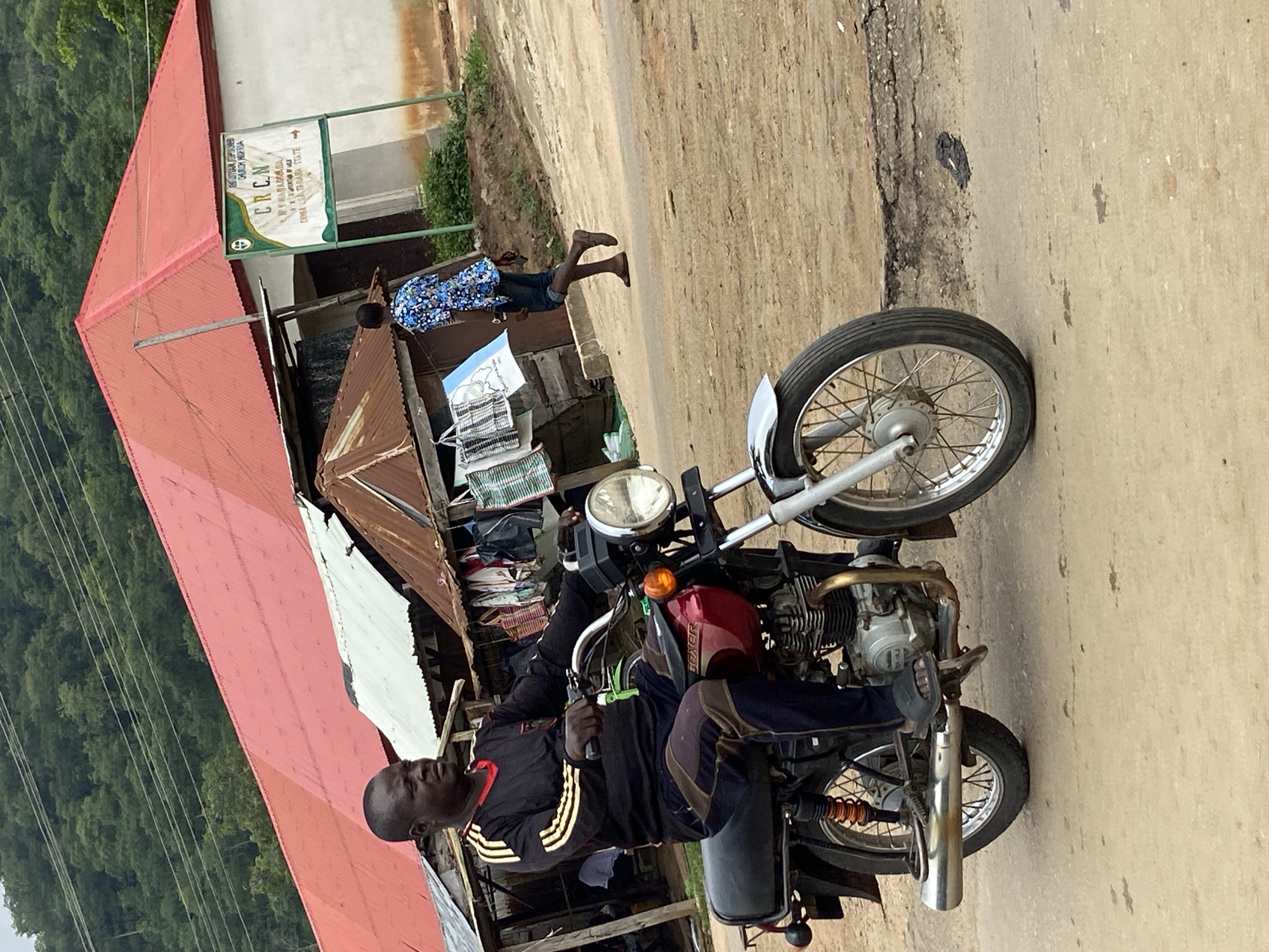 In Mararaba, Nigeria, a man rides a maroon motorcycle with a chrome exhaust pipe and silver spokes on a dirt road. He wears a black long-sleeved shirt with yellow stripes on the sleeves and dark pants with brown and yellow stripes. The motorcycle's headlight is on. In the background, a building with a red corrugated metal roof stands behind a row of small shops with tin roofs. One shop displays several bags for sale. A young person in a blue and white patterned shirt walks away from the shops. A sign on the building reads "ECUMENICAL REFORMED CHURCH NIGERIA" and "CRCN". The environment appears to be a rural or semi-urban area with vegetation on a hill in the distance.