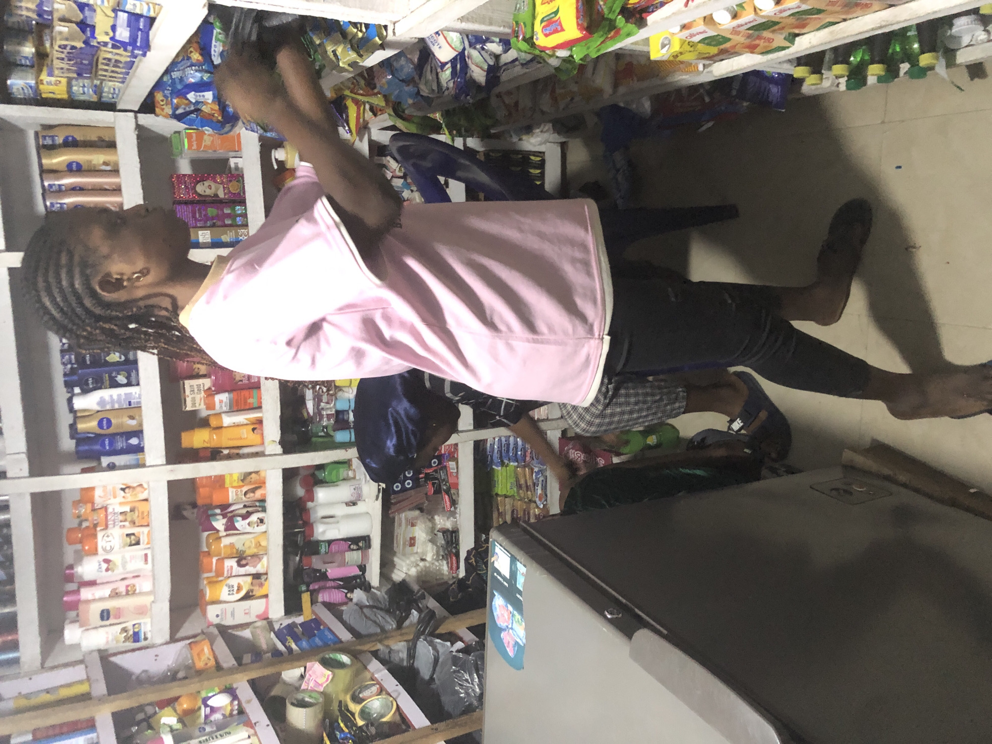 A young Black woman with braided hair is standing in a small shop in Ado, Nigeria. She is wearing a light pink t-shirt and dark leggings. She is reaching for items on a shelf. The shop is filled with shelves stocked with various products, including toiletries, packaged foods, and drinks. A refrigerator is visible to the left of the woman. Another person, wearing a dark head covering and checkered pants, is partially visible in the foreground, also appearing to be interacting with the shelves. The scene suggests a commercial environment with a focus on retail goods.