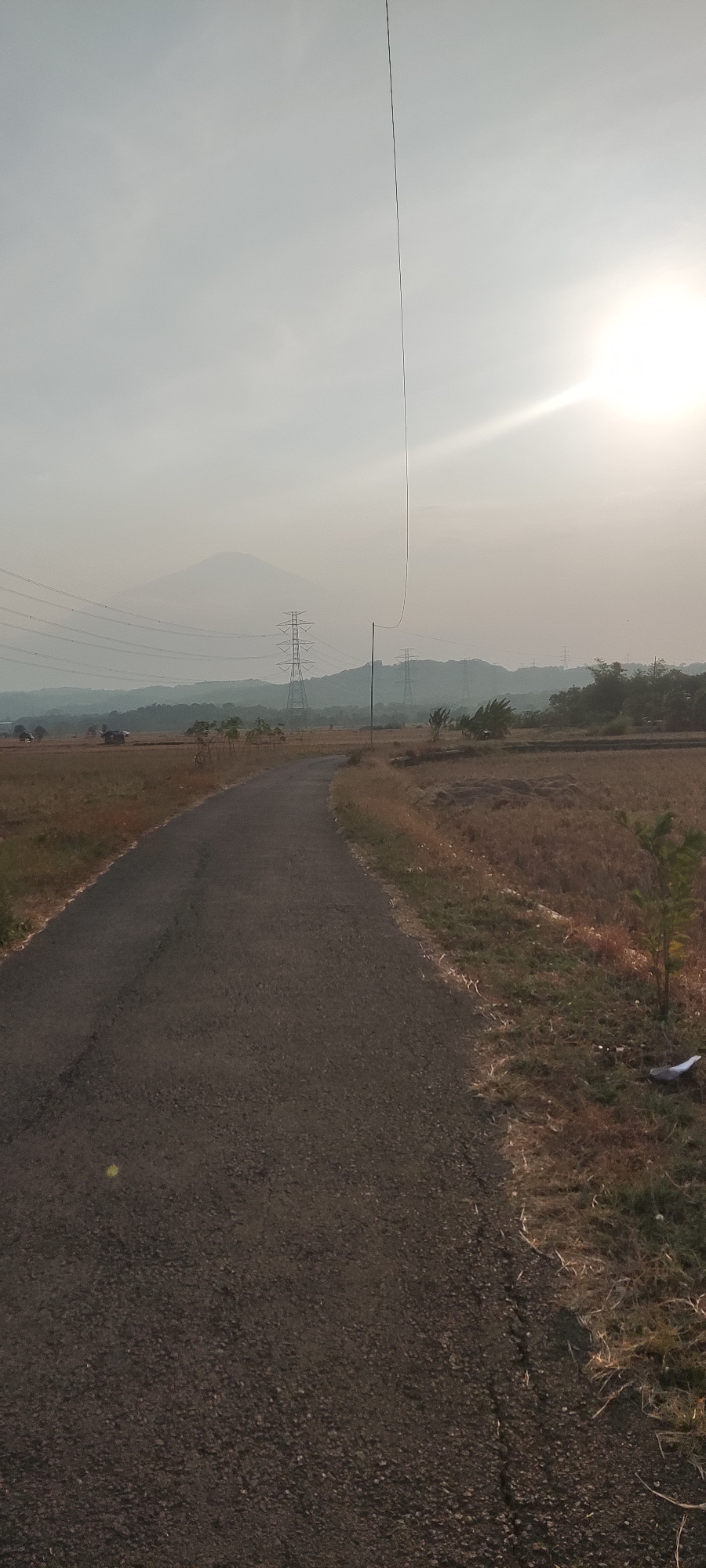 A paved road curves through a dry, grassy landscape. In the distance, hazy mountains are visible, with power lines and transmission towers dotting the terrain. A bright sun is setting in the upper right corner of the image, casting a warm glow over the scene. The overall impression is one of a rural area in Sedong Kidul, Indonesia, during either sunrise or sunset.