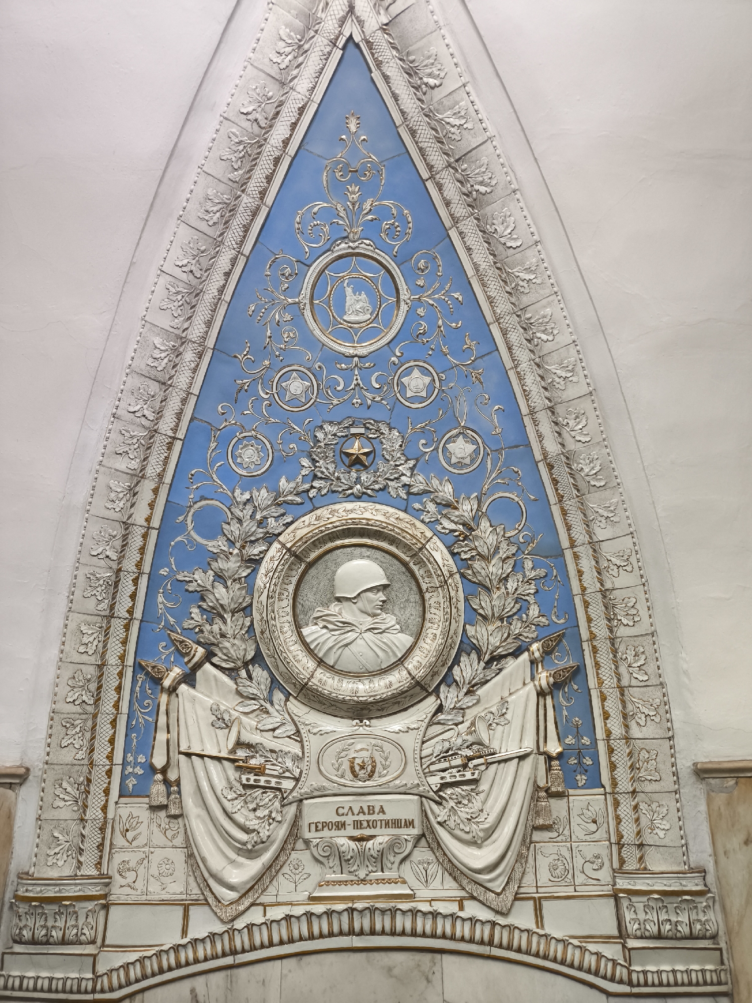 The image shows a large, triangular, bas-relief panel adorning a wall. The panel is primarily white and light blue, with gold accents. At the center is a circular medallion depicting a soldier in a helmet, facing right. Surrounding the medallion are laurel wreaths. Above the medallion are several circular emblems, including one with a star and others with abstract designs. Draped flags hang on either side of the medallion. Below the medallion is a plaque with the inscription "СЛАВА ГЕРОЯМ-ПЕХОТИНЦАМ" (Glory to the Heroic Infantrymen). The background is a light blue mosaic. The entire panel is framed by a decorative border of oak leaves and other ornamental designs. The wall surrounding the panel is a light beige color. The location is unknown.