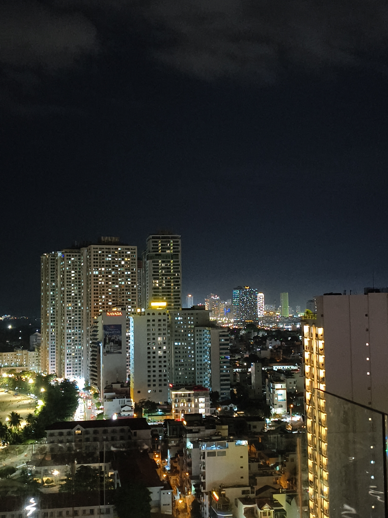A high-angle, full shot captures a city skyline at night. The sky is dark with visible clouds. In the foreground, a modern building with illuminated windows is on the right, partially obscuring the view. The midground features a cluster of tall buildings, many of which are lit from within, creating a grid of warm lights against the dark sky. One prominent building displays a banner that reads "NAVADA". The background reveals a sprawling urban landscape with numerous buildings and lights, suggesting a densely populated area. A street with cars can be seen at the lower left of the midground, and trees line the street. The overall scene depicts a vibrant cityscape at night.