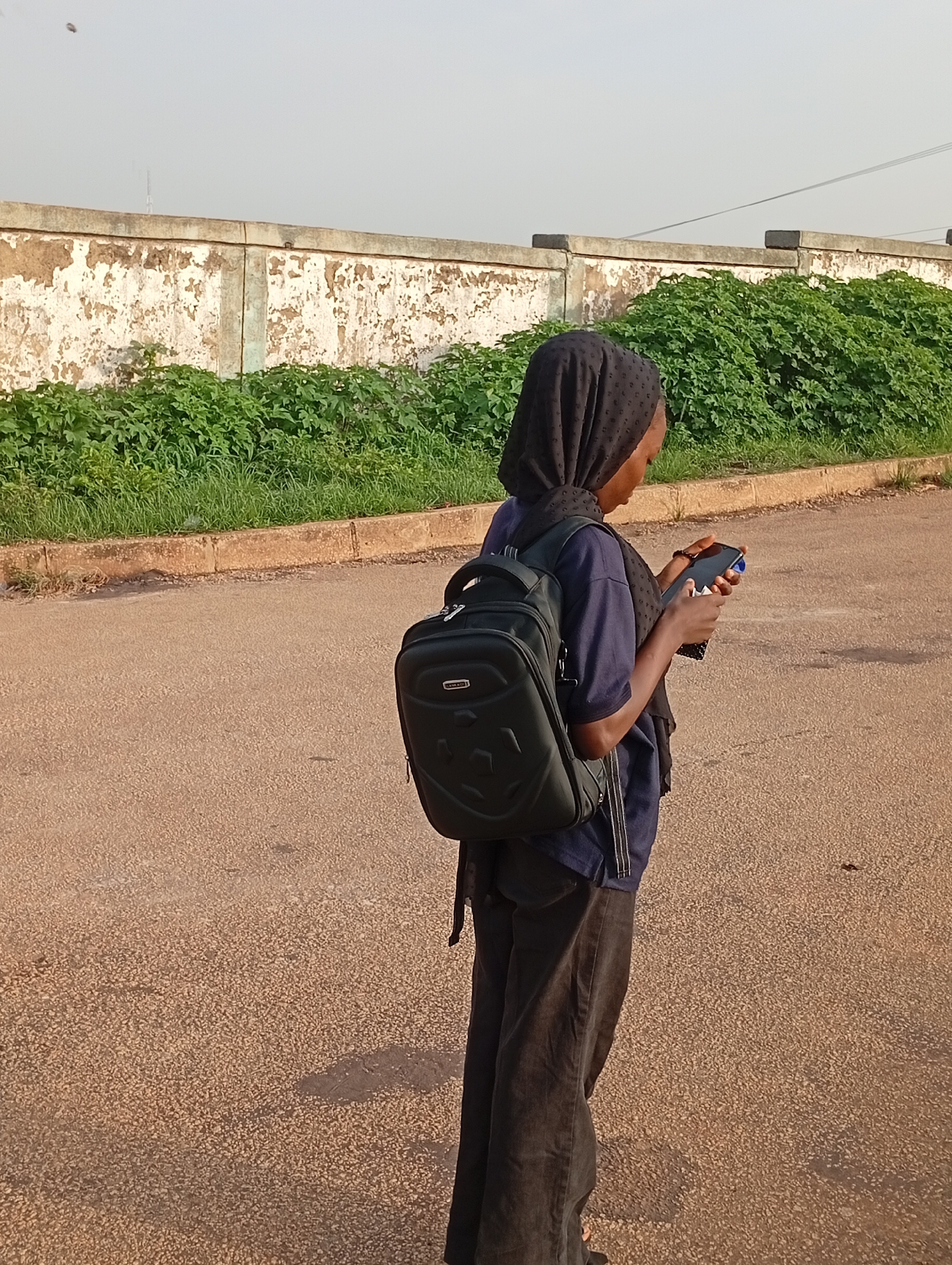 A young person wearing a dark blue t-shirt, dark pants, and a black hijab stands on an asphalt road, looking at a smartphone. They are carrying a black backpack with a silver zipper and a black logo. The person is positioned in the foreground, with a weathered concrete wall and a lush green hedge in the background. The wall appears to be crumbling in places, revealing patches of exposed concrete and dirt. The sky is a pale, hazy grey, suggesting it might be early morning or late afternoon. The ground is a reddish-brown asphalt, with some darker patches indicating moisture or debris. The overall setting appears to be an outdoor urban or semi-urban environment, potentially in a location like Rafin Par, Nigeria, given the visual cues.