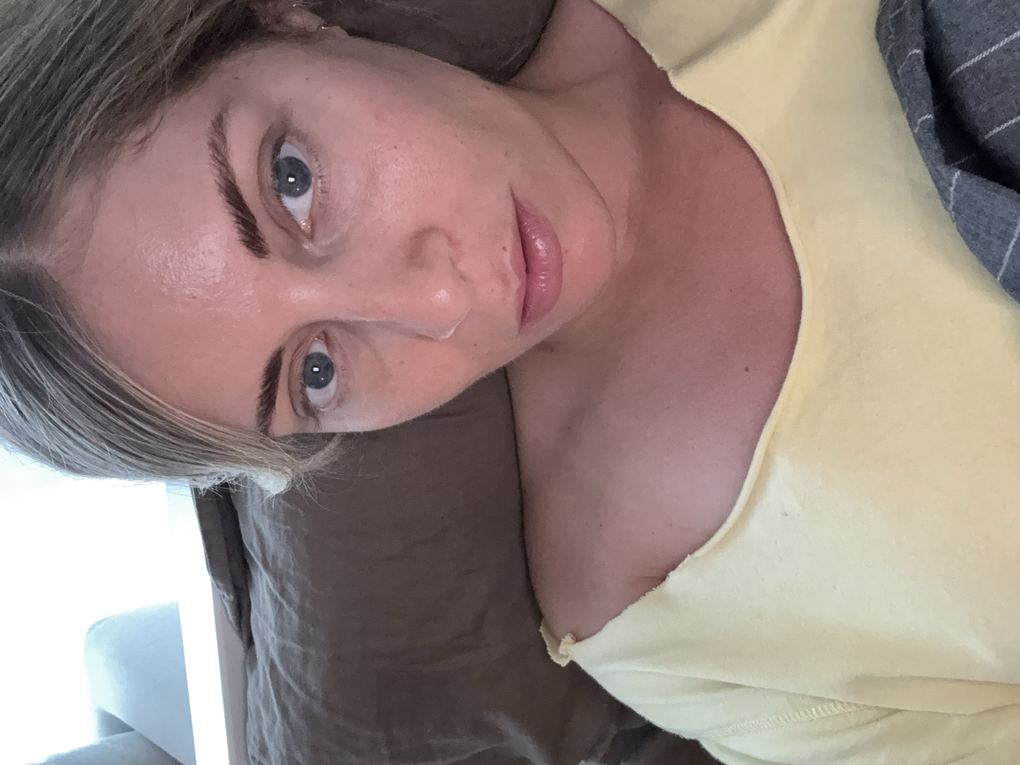 A woman with fair skin, light brown hair, and blue eyes is lying down. She is wearing a yellow top, and a gray plaid item is near her. The background includes a brown textured surface, possibly bedding, and a glimpse of a white object. There is no evidence of the city of Paris or France in the image.
