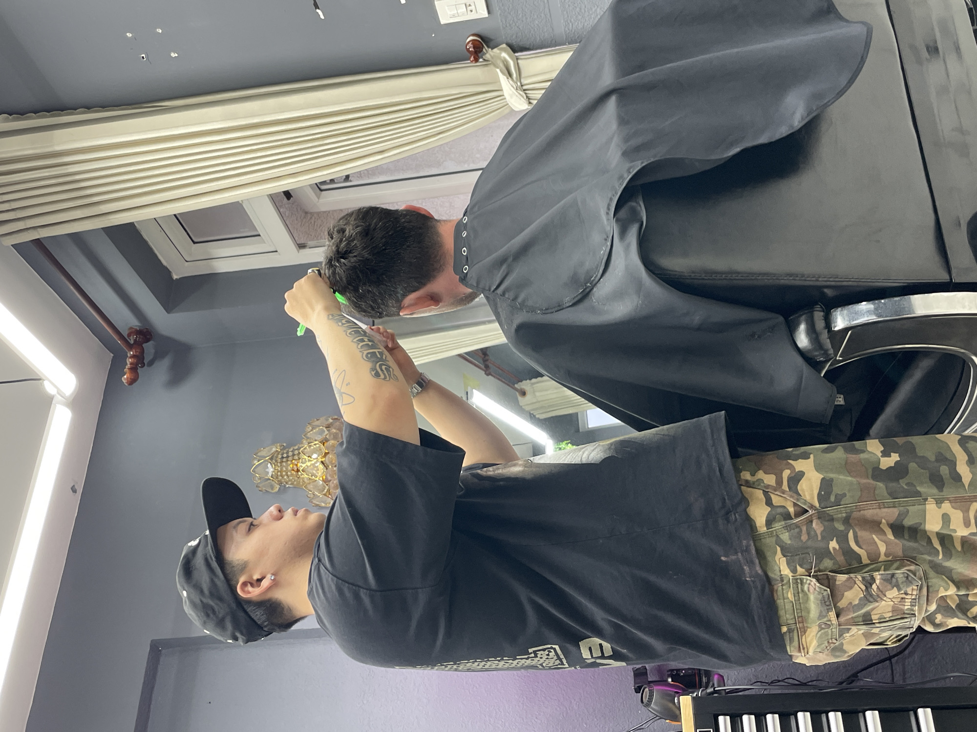 A barber is cutting the hair of a customer in a salon in Xuân Hương - Đà Lạt, Vietnam. The barber, wearing a black t-shirt and camouflage pants, stands to the left of the frame, holding scissors and a comb. The customer sits in a black barber chair, covered by a black cape. The background features a gray wall, a window with cream-colored curtains, and lighting fixtures. A decorative lamp and a tool cart are also visible.