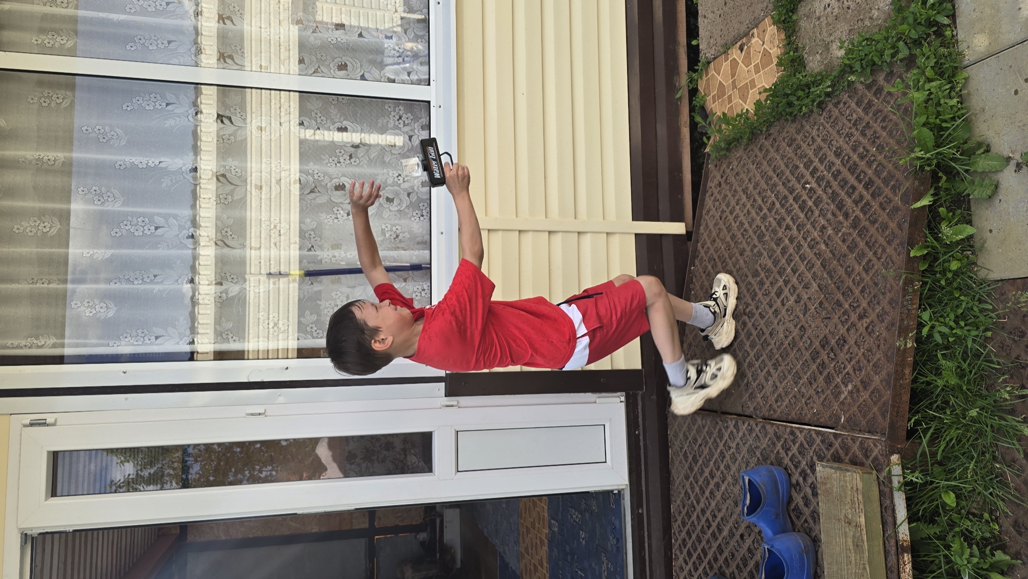In Забегалово, Russia, a young boy, aged approximately 7-10, is captured in mid-stride outdoors. He wears a red short-sleeved t-shirt, red shorts with a white hem stripe, white socks, and light-toned athletic shoes. He holds a black and clear plastic water gun, labeled "WATER GUN," in his extended right hand, aimed forward. His left arm is bent and extended with fingers splayed, and his facial expression suggests active engagement. He stands on a rusted metal diamond plate ramp. To the left foreground, a pair of blue slip-on shoes and a wooden block are visible on the ground. The background consists of a light beige building with horizontal siding, featuring white-framed windows displaying floral sheer curtains, and a partially open white-framed glass door. Green vegetation, including grass and weeds, grows around the ramp and adjacent concrete paving sections. The scene is illuminated by bright daylight.