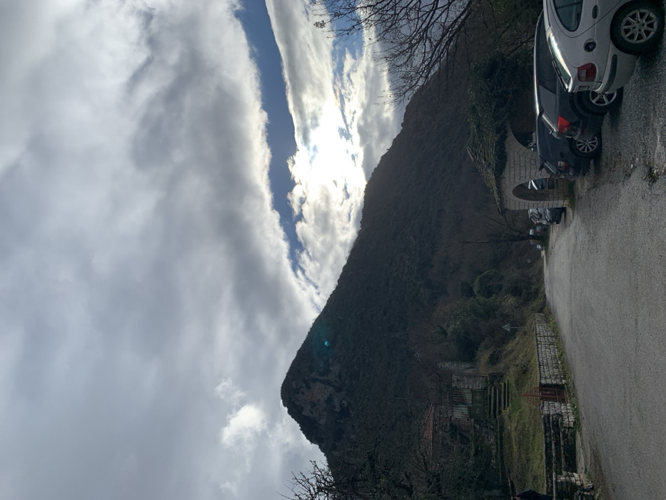 The image depicts a dramatic mountainous landscape in Vikos, Greece, under a bright, partially cloudy sky. A large, steep mountain, covered in dark, dense vegetation, dominates the right side of the frame, rising sharply towards the sky.
The sky above features a striking cloud formation: thick, grey clouds cover most of the upper area, but a distinct, elongated strip of clear blue sky is visible. The sun shines brightly through this opening, creating a strong light source and noticeable lens flare. The overall impression is a bright day with significant cloud cover. Bare tree branches are visible against the sky and along the mountain's lower slopes, suggesting late autumn or winter.
In the foreground, an asphalt road or path curves into the scene from the lower left towards the upper right. Several cars are parked along the side of this road, including a white and a dark-colored vehicle. Further along the road, an old stone archway or bridge structure is visible, integrated into the natural and built environment. Stone walls and what appear to be terraced, possibly disused, structures can be seen on the lower slopes of the mountain.
There are no people visible in the image, but the parked cars indicate recent or ongoing human activity in the vicinity. The time of day appears to be mid-day or early afternoon, characterized by the strong sunlight breaking through the clouds. No discernible text is visible on any objects or signs within the frame. The scene captures the rugged beauty of the Greek mountains alongside a quiet roadside setting.