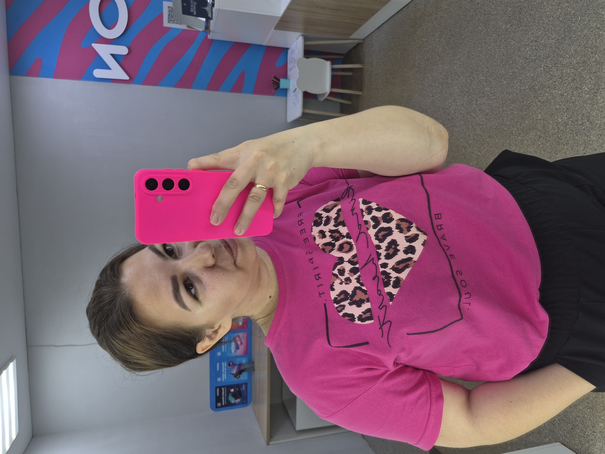 A woman is taking a selfie in a room with light-colored walls and a gray carpeted floor. She is wearing a bright pink t-shirt with a leopard print heart design and black pants. She is holding a pink smartphone in her right hand, and a gold ring is visible on her left ring finger. In the background, there is a wall with a pink and blue striped pattern and white lettering that reads "МОЙ" (MY in Russian). To the right, a small white table with a white chair shaped like a bear's head is visible. The scene suggests an indoor retail or service establishment. The presence of Russian lettering indicates the location is in Russia. Given the context of a store with signage that includes Russian text and the overall interior design, the location is likely a retail or service point within the city of Chaikovsky, Russia.