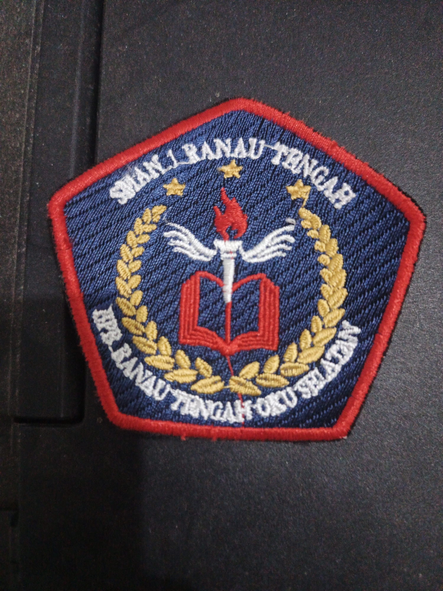 An embroidered patch with a pentagonal shape and a red border is displayed on a dark, textured surface. The patch is primarily dark blue with white and gold embroidery. At the top of the patch, white text reads "SMAN 1 RANAU TENGAH". Below this, a gold laurel wreath surrounds a central emblem. The emblem features a stylized open red book with a red flame atop a white torch. On the lower portion of the patch, white text follows the curve of the laurel wreath and reads "APR. RANAU TENGAH OKU SELATAN". The overall design appears to be a school or organizational crest. The dark background suggests the patch is lying on a surface such as fabric or a similar material. There is no indication of a specific city or location within Indonesia.