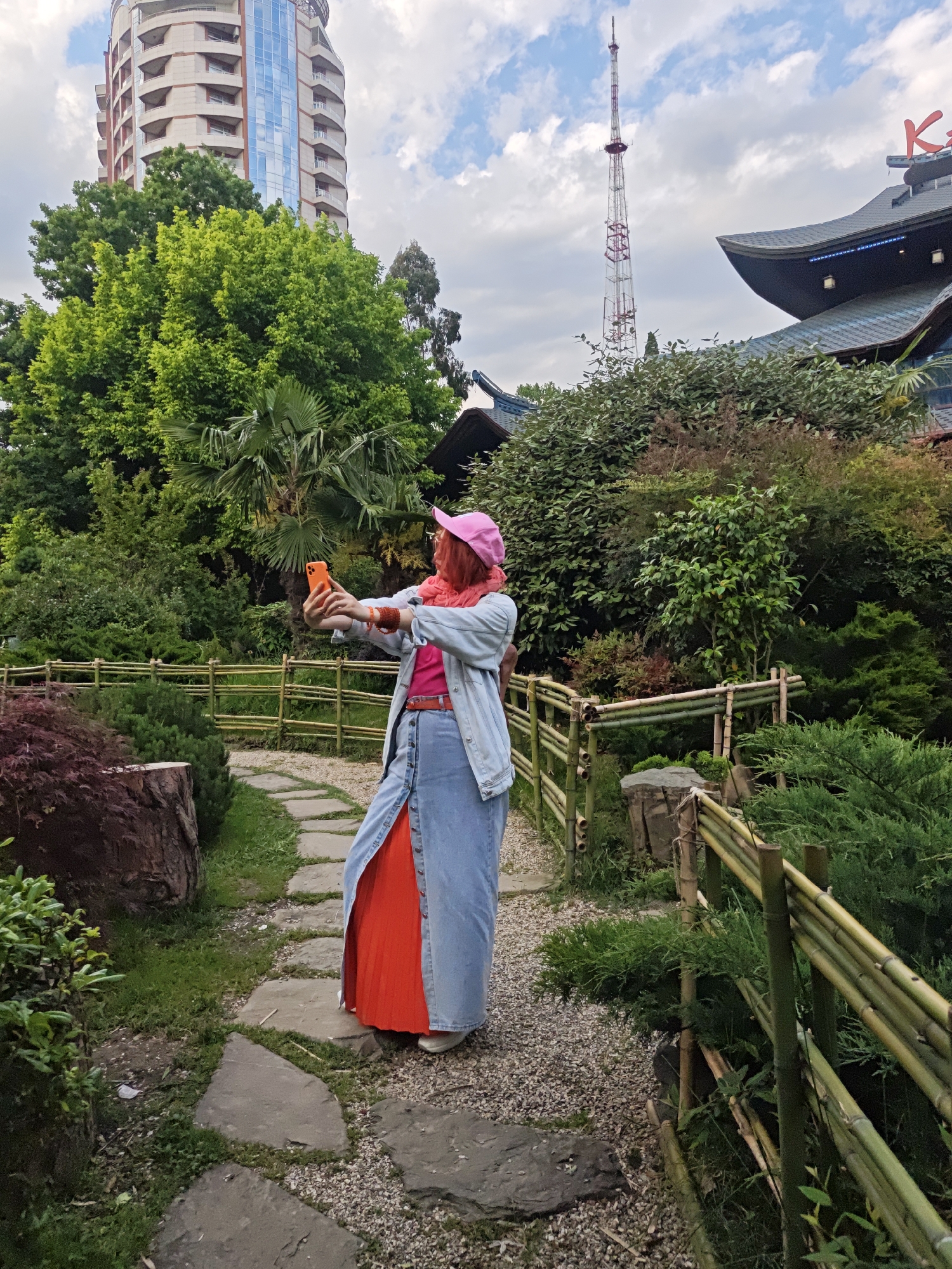 A full-length shot captures a woman in a brightly colored outfit taking a selfie in a lush garden. The woman is facing away from the camera and towards a pathway in the garden. She is wearing a pink cap, a pink top, a light blue denim jacket, and a long blue skirt with a vibrant orange under-skirt. Her right arm is extended, holding an orange mobile phone. A red belt is visible around her waist.
The garden has a stone pathway lined by low bamboo fences and various greenery, including shrubs and trees. In the background, there is a tall red and white communications tower, a building with architectural designs similar to a Japanese temple, and a modern high-rise building.
This scene is set in the Garden of Russian-Japanese Friendship in Sochi, Russia.