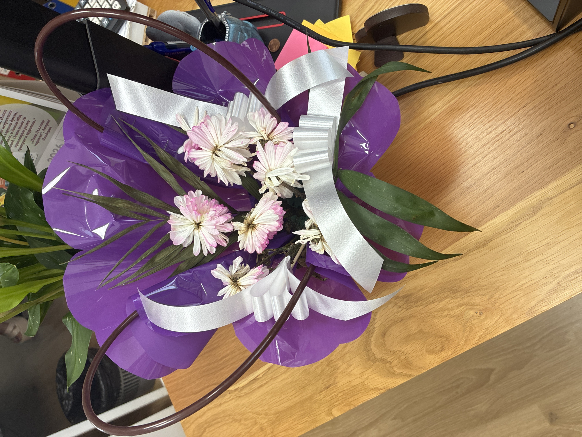 A heart-shaped bouquet of flowers is displayed on a wooden table. The bouquet is wrapped in purple paper and decorated with white ribbons and bows. Several light pink and white daisy-like flowers are visible, along with some green foliage. A dark brown wire frame forms the heart shape around the bouquet. In the background, there are plants, papers, office supplies, and a black computer monitor. The table appears to be made of light-colored wood with distinct grain patterns.