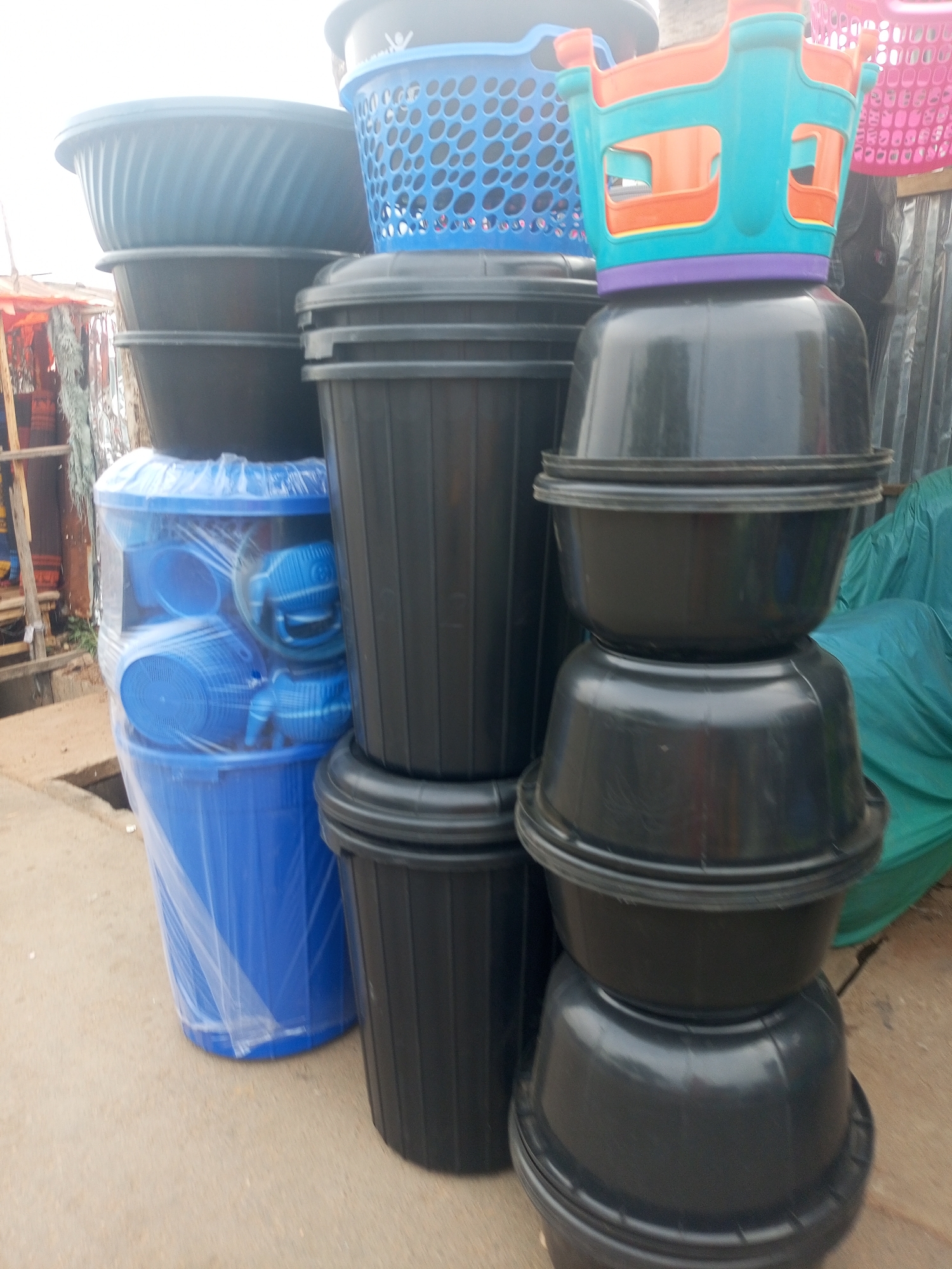 The image depicts a roadside display of plastic containers in Gombe, Nigeria. There are several stacks of containers, including blue plastic wash basins wrapped in clear plastic, black cylindrical containers, and black rounded basins stacked on top of each other. A blue and orange plastic basket sits atop the rounded basins. The scene appears to be in a market area.
