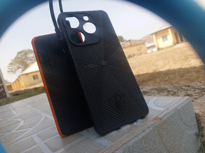 The image displays two smartphone cases positioned outdoors in Kuta, Nigeria. A black textured smartphone case, featuring cutouts for a multi-lens camera array and a geometric pattern, stands upright in the foreground. A dark cable connects to its upper left side. Behind it, a second dark-colored case with an orange perimeter trim is partially visible, displaying partial text on its surface. These items rest on a light-colored, tiled surface, likely a concrete ledge or block. The background is out of focus, revealing dry, light-brown ground, several blurry single-story buildings with light-colored walls and dark roofs, and a sparse tree on the left. The sky is clear and light blue, indicating bright daylight conditions. A blue curved artifact partially frames the top and bottom edges of the image. No individuals are present in the scene.