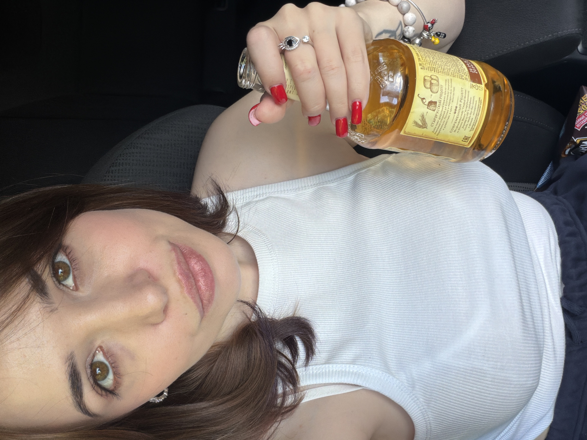 A person lies on their back, with their head tilted and turned to the side, looking directly at the camera. They have long, dark brown hair, fair skin, and light brown eyes. They are wearing a white tank top and a dark blue garment underneath. Their right hand, with bright red nail polish, holds a glass bottle with a yellow liquid inside, labeled in Russian with "СТАРОРУССКИЙ Рецепт" (Old Russian Recipe) and other text. A silver ring with a dark stone and a smaller clear stone is worn on their index finger, and they have a bracelet with several charms on their wrist. The setting appears to be indoors, possibly in a car, given the dark background and textured dark material. The lighting suggests it could be daytime. The person's expression is neutral.