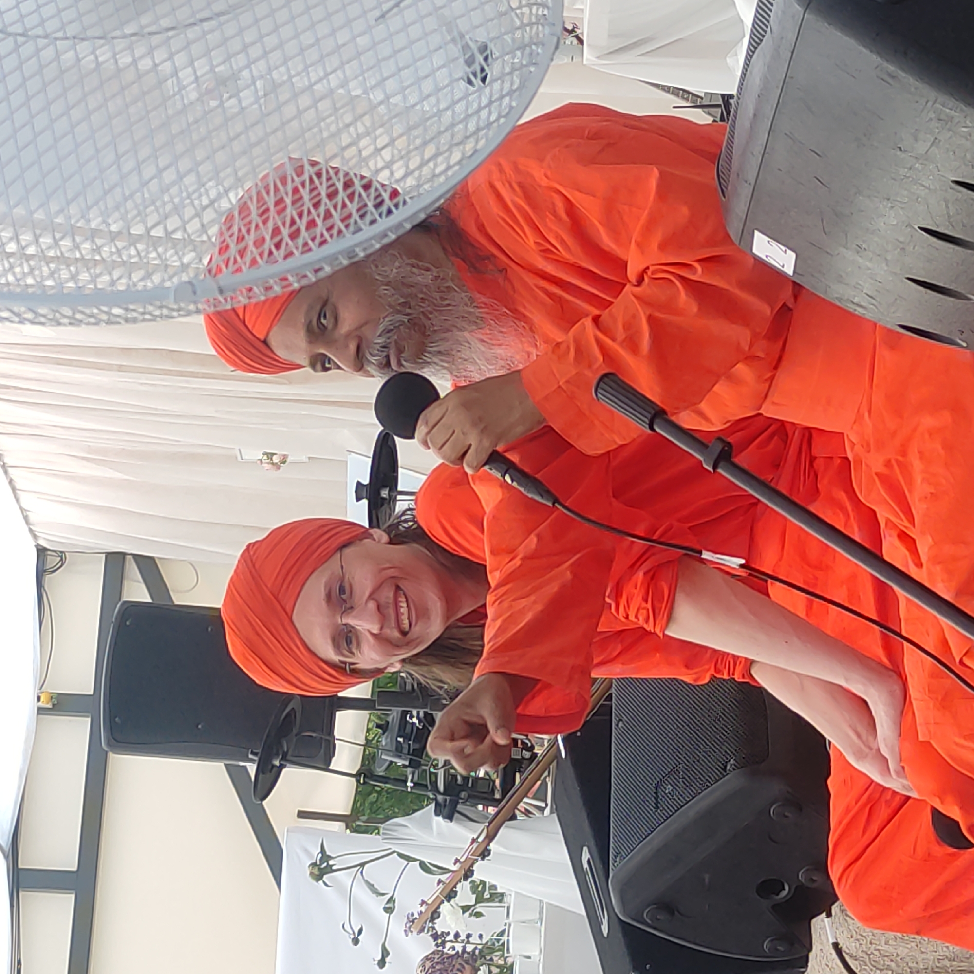 Two individuals, both wearing orange robes and turbans, are seated on a stage outdoors. The person on the left, with glasses and long brown hair, is smiling and pointing towards the viewer with their right index finger. The person on the right, with a gray beard and mustache, is holding a microphone to their mouth and appears to be speaking. They are positioned behind a microphone stand. In the background, a white curtain and a large speaker are visible. A white, mesh-like object, possibly a fan or decorative screen, is in the foreground on the right, partially obscuring the person on the right. The event appears to be a public gathering or performance. The location is identified as Zhirnosovo, Russia.