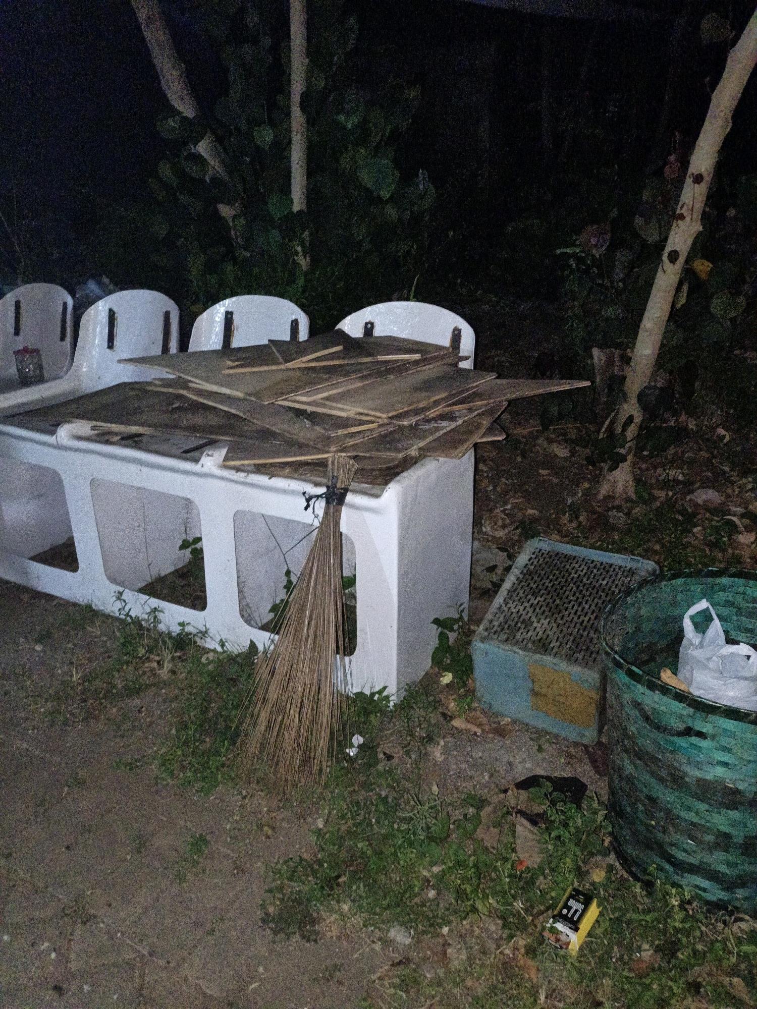 The image depicts an outdoor scene at night, possibly in Cikal, Indonesia, with a focus on a white, bench-like structure laden with pieces of wood. To the right of the bench, a broom made of dried leaves is leaning against it. Further to the right, a blue and yellow crate-like object is visible, next to a large, green, woven basket containing a white plastic bag. The ground is covered with dry grass and dirt. Trees and foliage form a dark backdrop, suggesting a natural or semi-natural environment. The lighting is dim, and the overall impression is one of disarray or temporary abandonment.