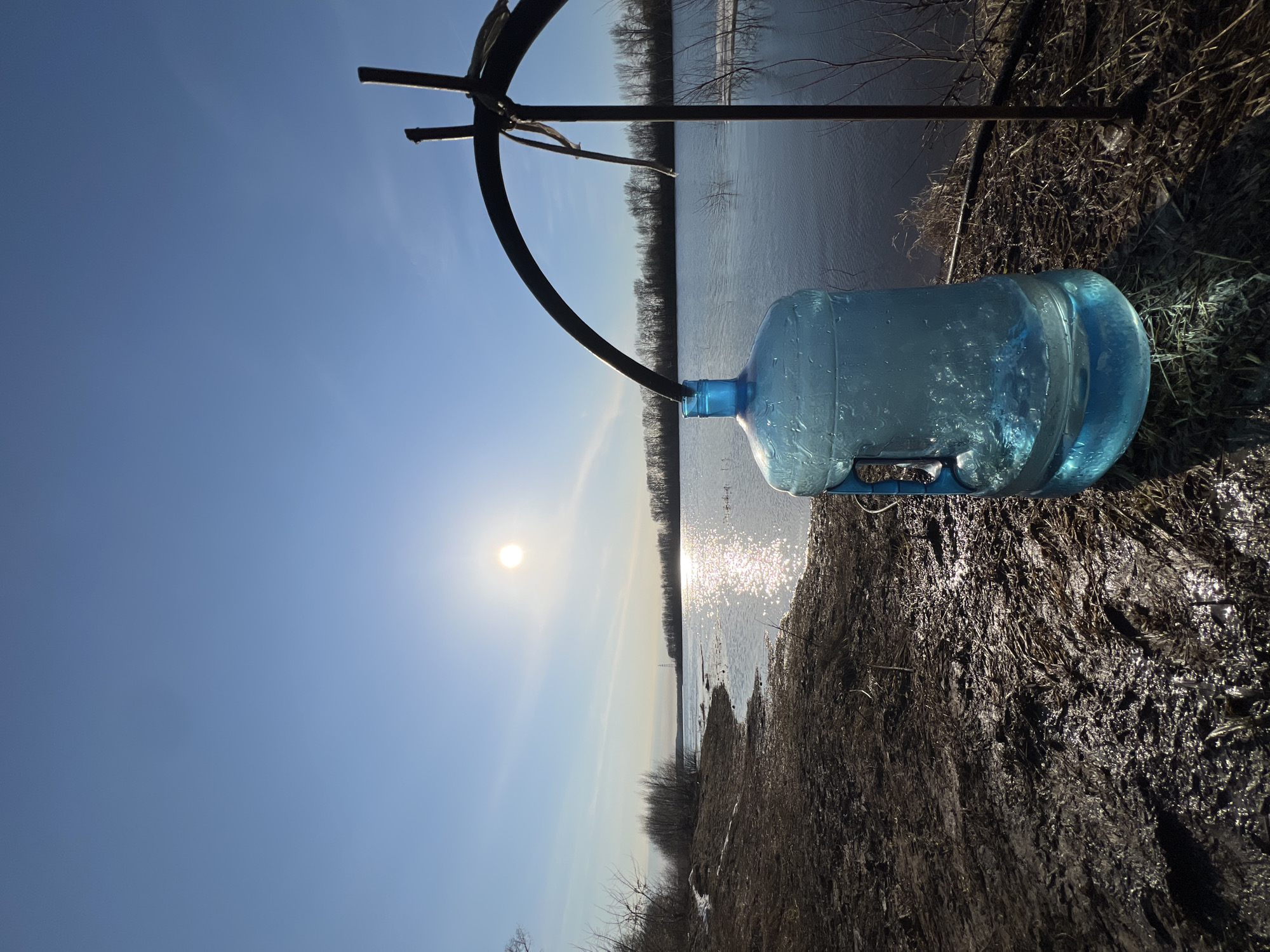 The image shows a 5-gallon blue water bottle lying on the ground near a body of water. A black metal frame is positioned above the bottle. The ground is muddy and covered with dead grass. In the background is a river or lake reflecting sunlight, with trees lining the opposite shore. The sky is blue with the sun visible. The location may be near a body of water in Chaikovsky, Russia.