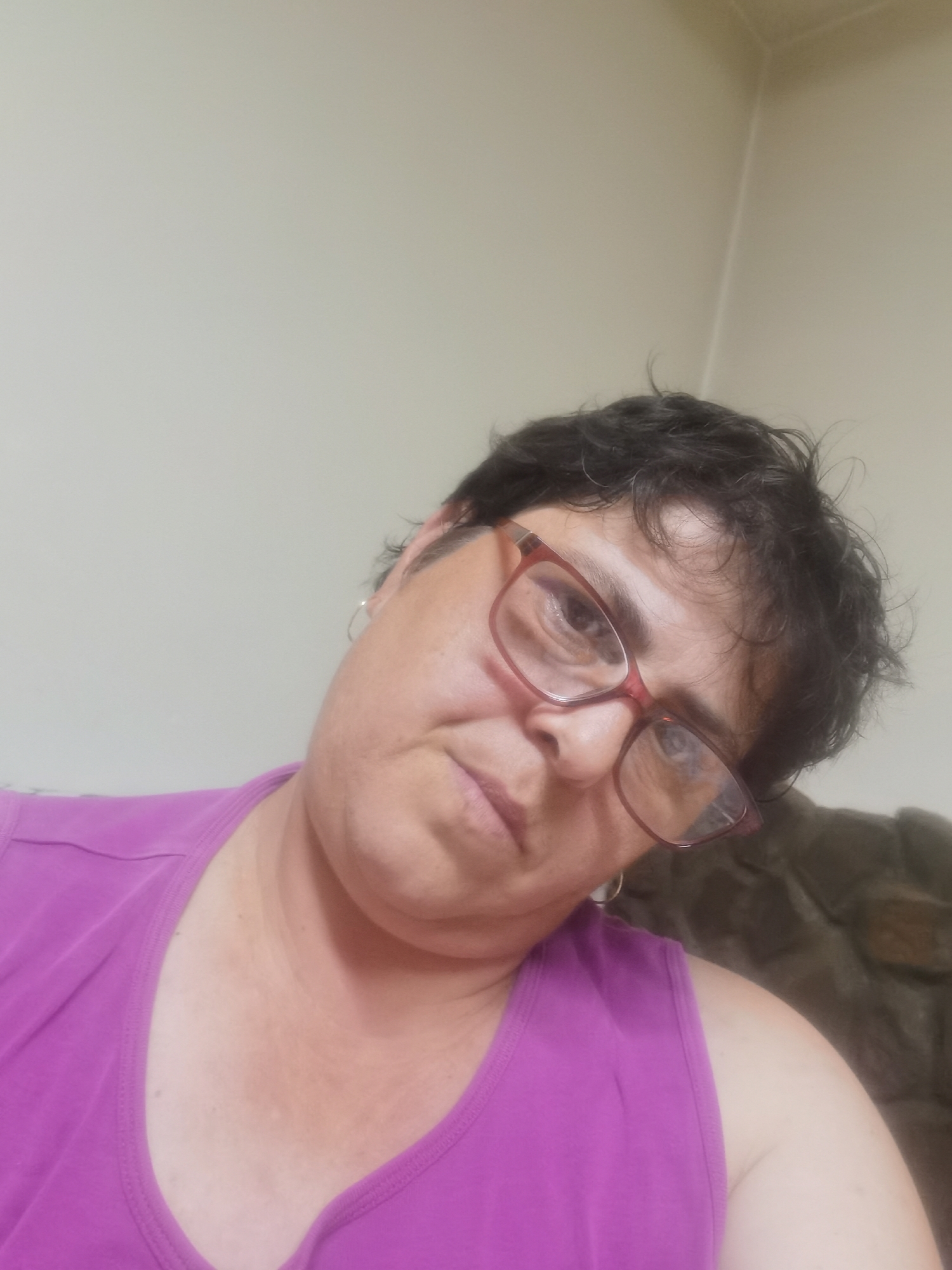 An individual is depicted in a head-and-shoulders selfie, captured indoors against a light-colored wall. The subject, appearing to be an adult, wears dark, short, and somewhat disheveled hair. They are adorned with red-framed eyeglasses and a small, circular earring in their visible left ear. The individual's head is tilted slightly to the left, and they gaze directly into the camera with a neutral expression. They are dressed in a bright magenta or fuchsia-colored sleeveless top with a scoop neckline. The background consists primarily of a plain, light-toned wall, with a corner visible in the upper right. A dark, textured object, possibly furniture or a decorative element, is partially visible on the far right behind the subject's right shoulder. The specific location in the city of Lomets, Bulgaria, is not discernible from the visual information provided in the image.