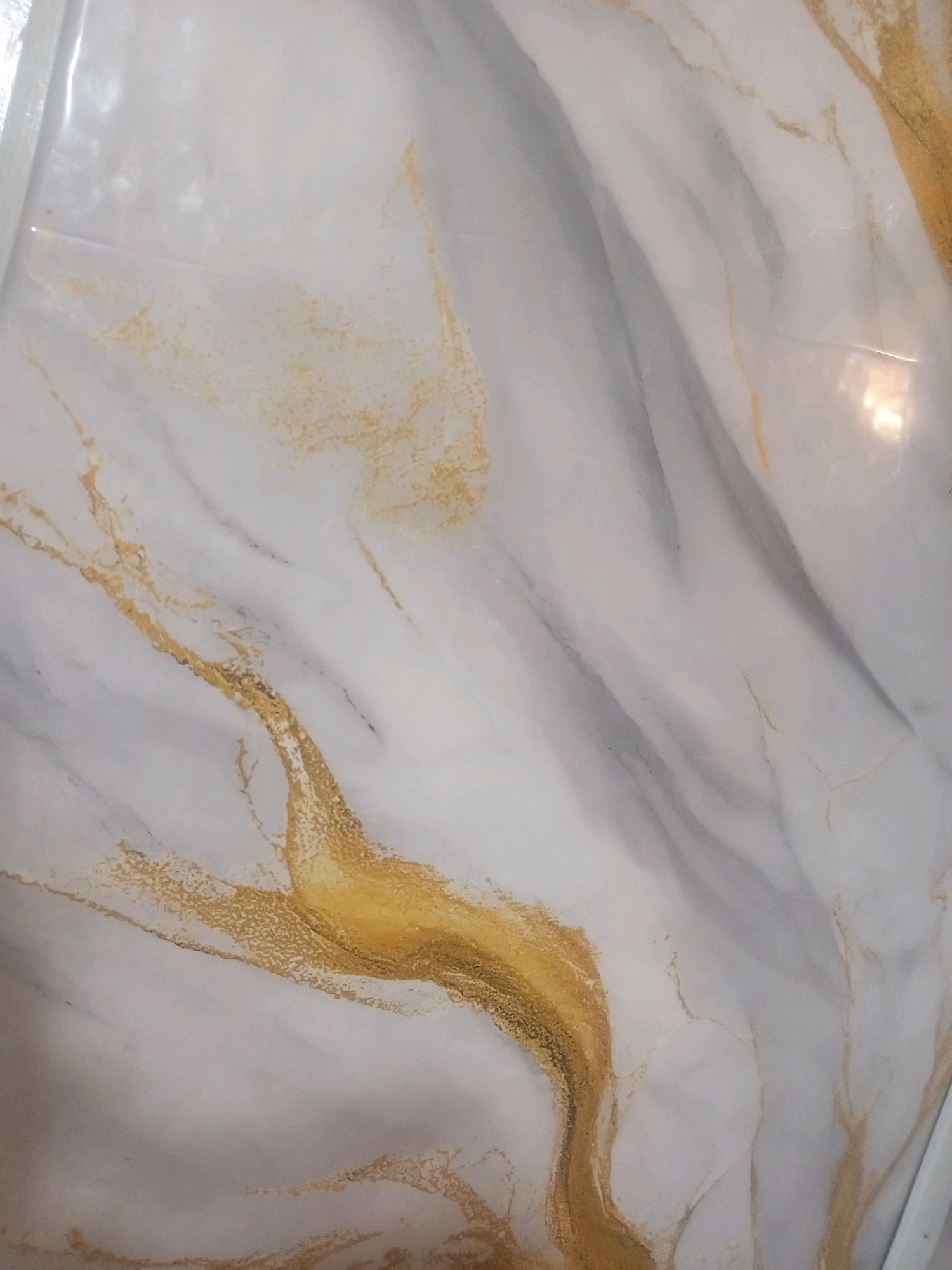 The image displays a close-up view of a marble-like surface with white, gray, and gold veining. The texture appears smooth and polished. The lighting suggests an indoor environment with reflections on the surface. There are no people, animals, or identifiable objects other than the material itself. The location in Jardy-Su, Kyrgyzstan, cannot be determined from the image.