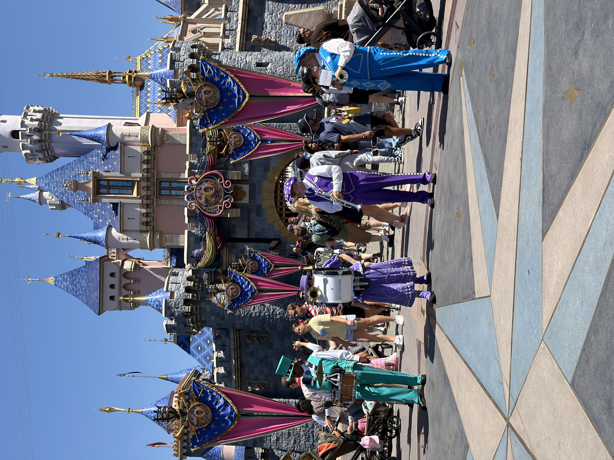 This image captures a bright, sunny day at Disneyland in Anaheim, California. In the foreground, a group of performers dressed in colorful marching band uniforms stand on a patterned tiled plaza. One performer in a teal suit plays a drum set, while others in purple and blue outfits play brass instruments. Behind them, the iconic Sleeping Beauty Castle is visible, adorned with festive banners celebrating the "70"th anniversary. A crowd of park-goers, including families and children, are seen walking and observing the performers. Strollers and shopping bags are also present, indicating a typical theme park environment. The overall atmosphere appears lively and celebratory.