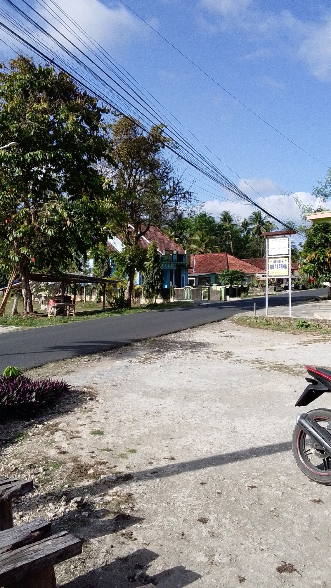 A motorcycle is parked on the right side of the image, facing away from the viewer. The motorcycle's rear wheel and part of its body are visible. The foreground consists of a concrete area with scattered debris and shadows. A wooden bench is partially visible on the lower left. In the mid-ground, a paved road curves to the left, bordered by a patch of dark purple foliage on the left and a sidewalk on the right. Along the road, several buildings with red-tiled roofs are visible, set back from the road and surrounded by trees. One building is painted blue and green, another is primarily red. A signpost is located on the right side of the road, with text that appears to be in Indonesian, including the word "APOTEK." The background features lush green trees and a clear blue sky with scattered white clouds. The scene depicts a street in a village or suburban area, likely in Cintaratu, Indonesia, given the architecture and vegetation.