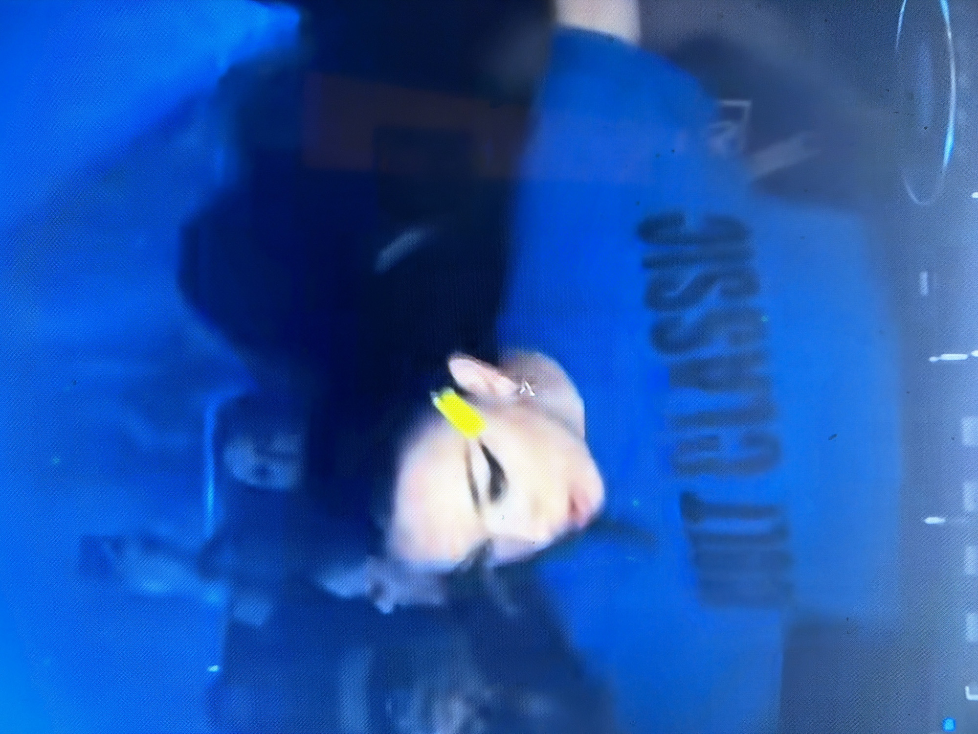 An indoor scene, likely a club or event venue in Maidstone, United Kingdom, is captured under dominant blue lighting, creating a dimly lit and atmospheric environment. In the central foreground, a person is visible from the chest up, positioned behind what appears to be performance or DJ equipment, with their head slightly tilted downwards and mouth slightly ajar, indicating focus or engagement. This individual has dark hair pulled back, prominent black winged eyeliner, and a distinct bright yellow hair accessory positioned on the right side of their head, alongside a silver-colored earring in their right ear. They are wearing a vibrant blue t-shirt emblazoned with the black text "CULT CLASSIC" horizontally across the chest. In the blurred background, multiple indistinct figures are present, some appearing to hold up mobile devices, suggesting an audience or gathering observing the central figure in the low light conditions. The overall image has a grainy texture, consistent with low-light photography.