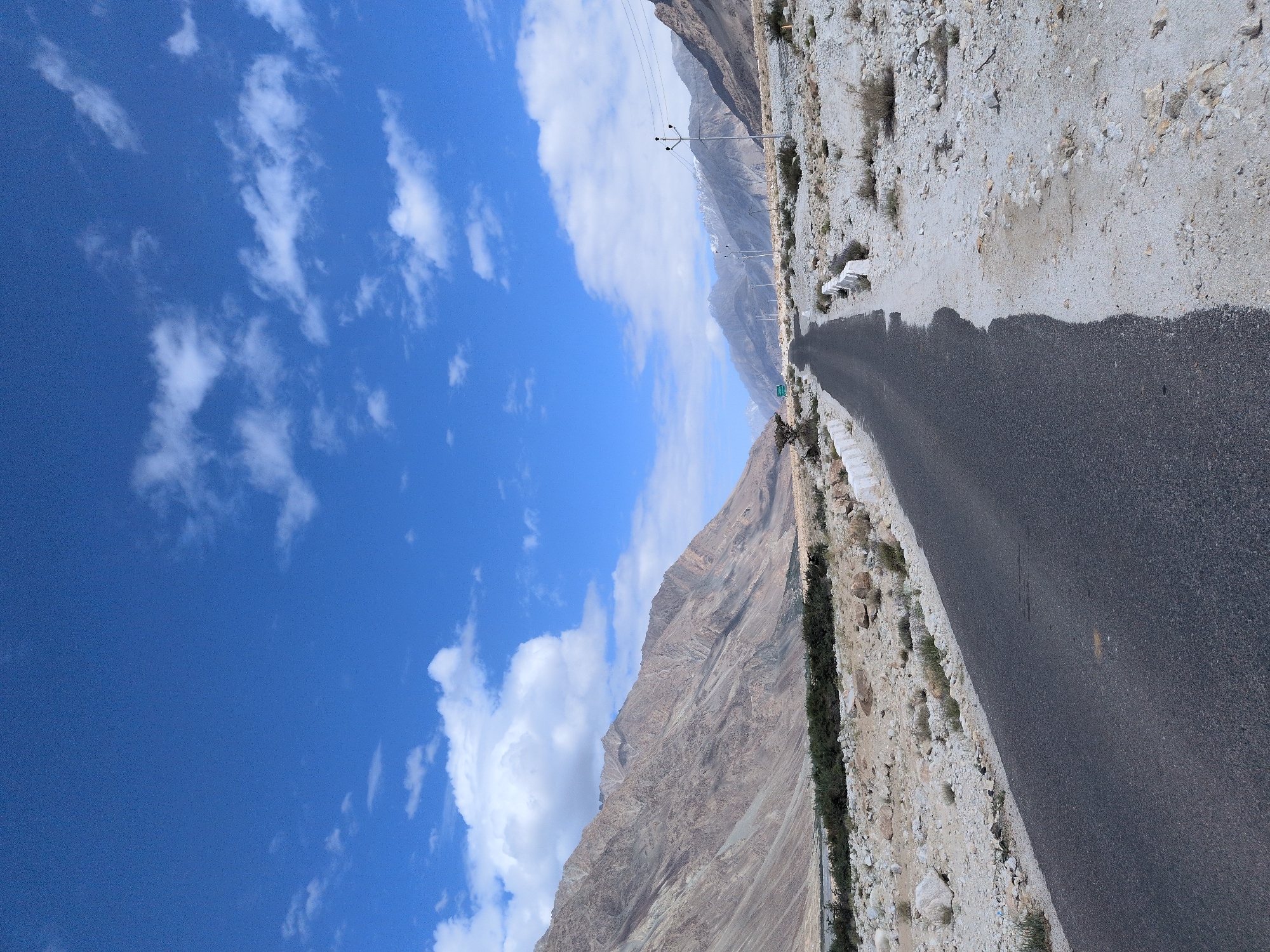 This is a photograph of a desolate road in Sasoma, India, under a bright blue sky with scattered white clouds. The road itself is asphalt and curves into the distance, flanked by a dry, rocky landscape. On the left side of the road, a rugged, barren mountain range rises sharply, its slopes showing layers of rock and sparse vegetation. On the right side of the road, the terrain is flat and covered with pebbles and rocks, with a few hardy bushes. In the distance, more mountains are visible, some with snow-capped peaks. Several utility poles with wires are scattered along the right side of the road, indicating some form of infrastructure. The overall scene is one of vastness and isolation. There are no people or animals visible in the image.