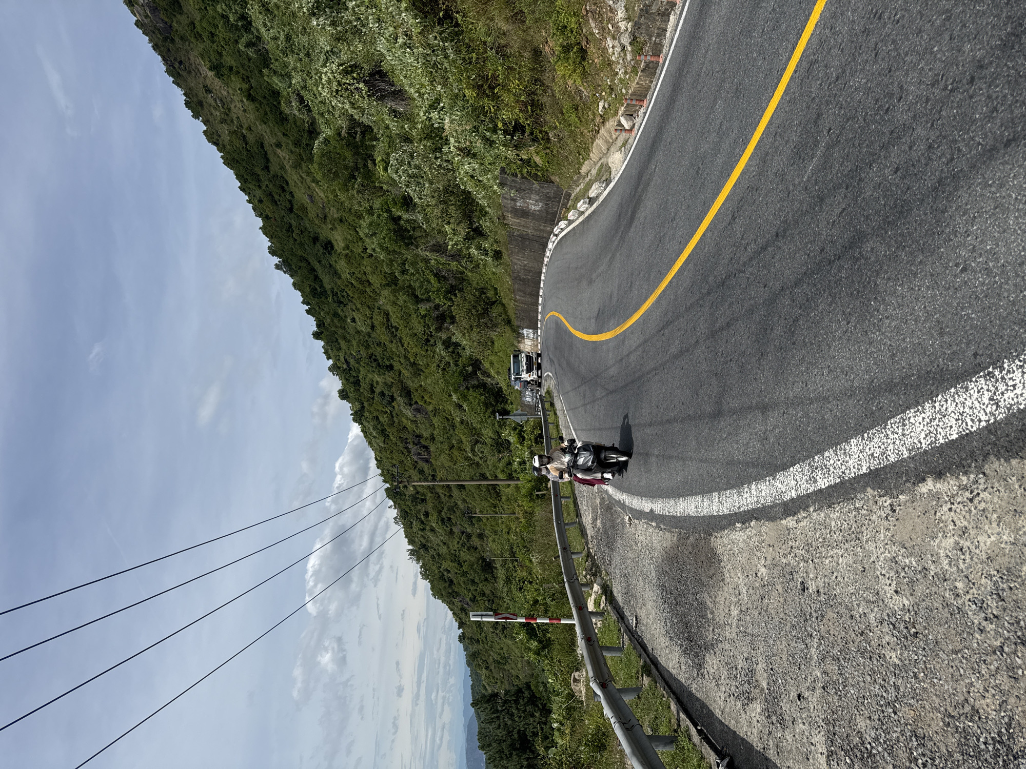 The image depicts a winding mountain road in Đà Nẵng, Vietnam, likely part of the Hải Vân Pass. A motorcycle carrying two individuals, both wearing helmets and one with a red scarf, travels along the asphalt road, which is marked with a white dashed line on the left and a solid yellow line down the center. The road is bordered by a metal guardrail on the left and a concrete retaining wall on the right, supporting a lush, green mountainside covered in dense vegetation. Overhead, power lines stretch across the sky, which is partly cloudy. In the distance, hazy mountains are visible. The scene is captured from a perspective slightly above and behind the motorcycle.