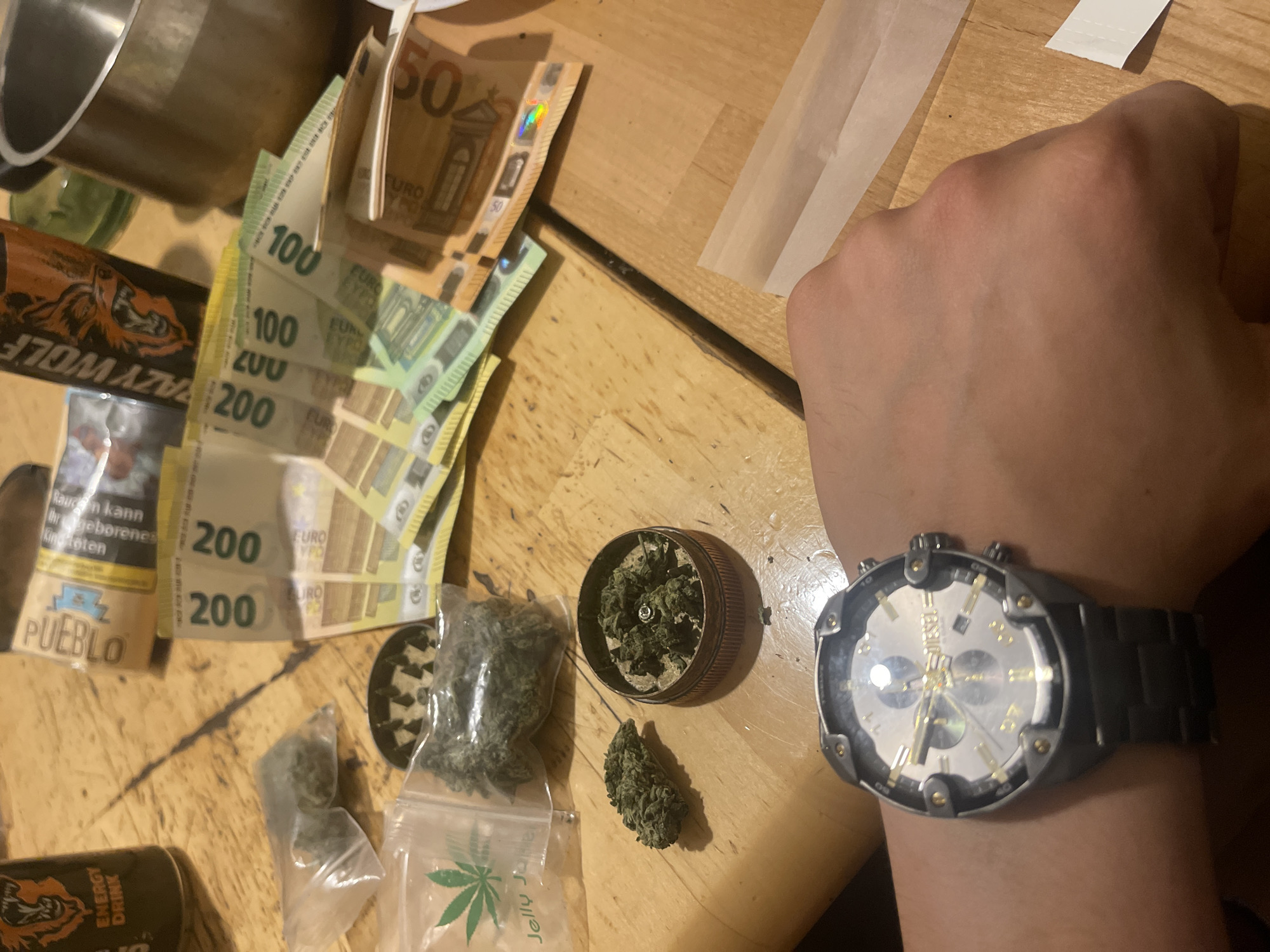 The image shows a tabletop scene with cannabis-related items, currency, and personal items. A hand wearing a Diesel wristwatch is visible in the lower right of the image. Next to the watch are cannabis buds, a grinder filled with ground cannabis, and a small plastic bag with a cannabis leaf design, also containing cannabis. A pile of Euro currency, including 50, 100, and 200 Euro notes, is arranged near the center. Other items include a can of "Lazy Wolf" energy drink, a pack of Pueblo rolling tobacco, and rolling papers.