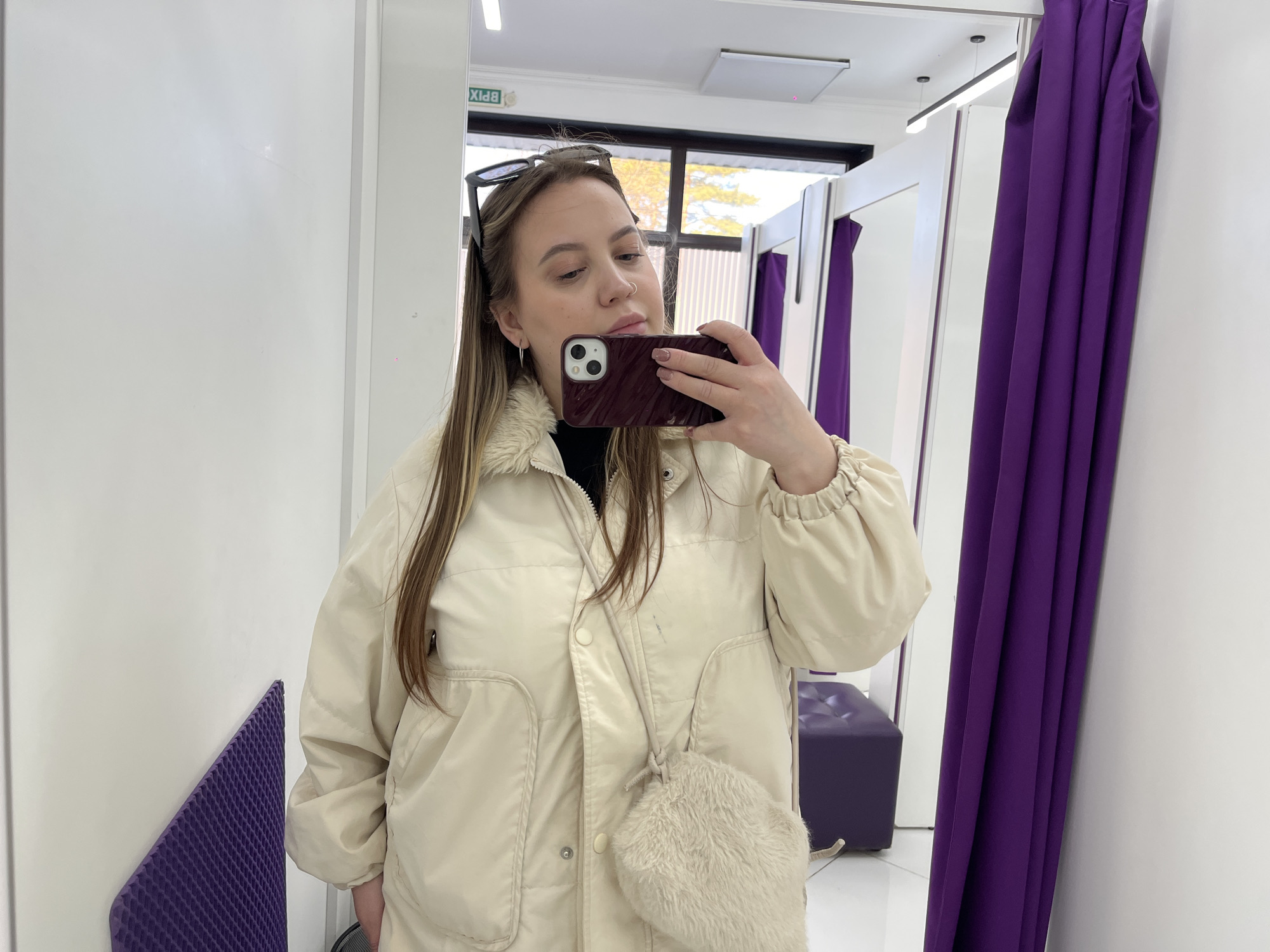 A woman is taking a selfie in a dressing room. She is wearing a cream-colored jacket with a faux fur shoulder bag, and her sunglasses are perched on her head. The dressing room has white walls, purple curtains, and a purple stool. There is a window in the background. A sign in the dressing room is in Russian, indicating the location is Novopokrovskaya, Russia.