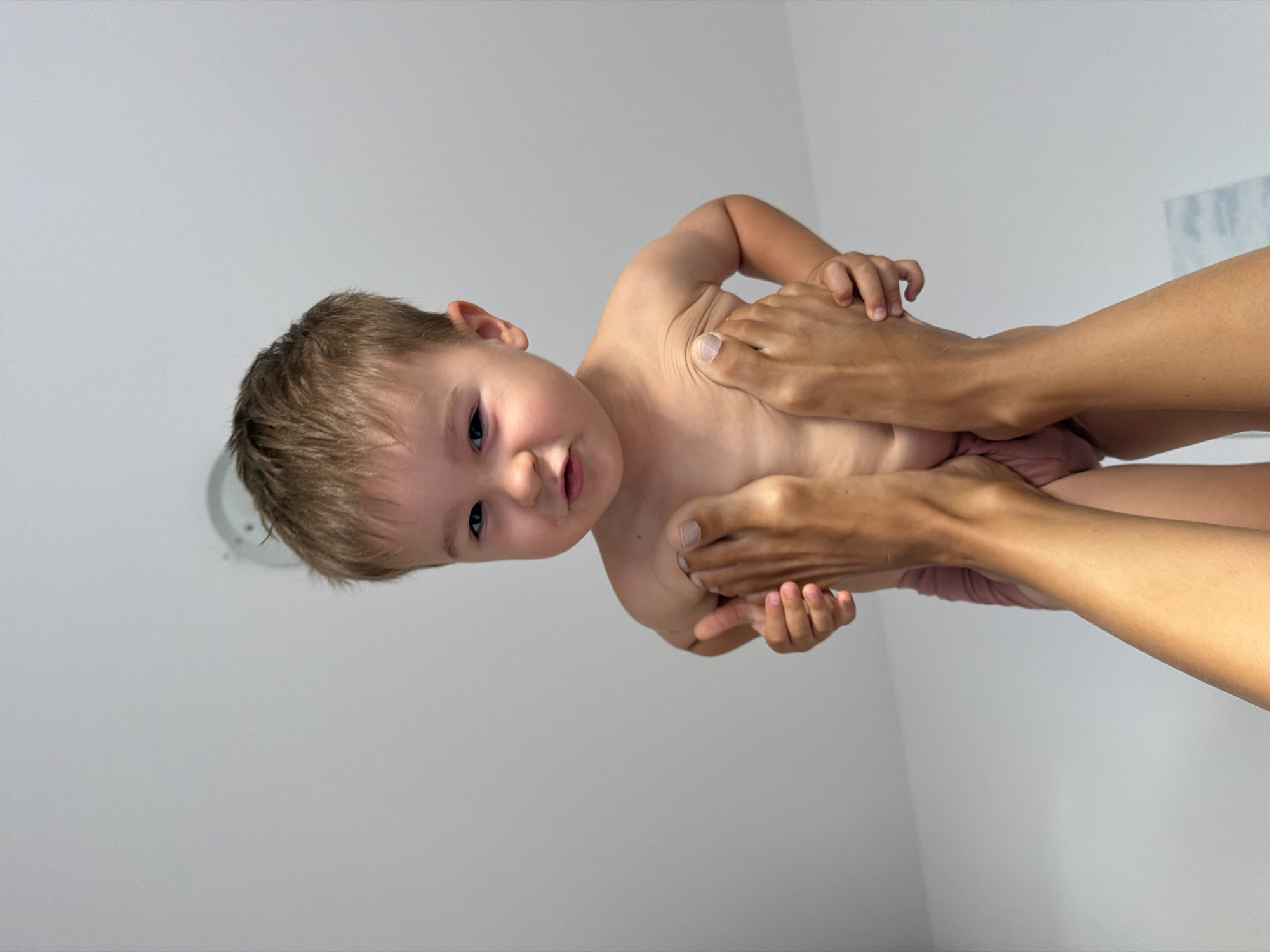 The image shows a close-up, vertically oriented shot of a young child being held by an adult. The child, who appears to be a toddler, is naked and has light brown hair and blue eyes. The adult's hands are visible, gently supporting the child's torso. The background is a plain, light gray wall, with a ceiling light fixture visible in the upper left corner. The lighting is bright, suggesting it is daytime. The child's expression is neutral, and the overall scene appears calm and intimate. The image was taken in Budva, Montenegro. There is no visible text in the image.