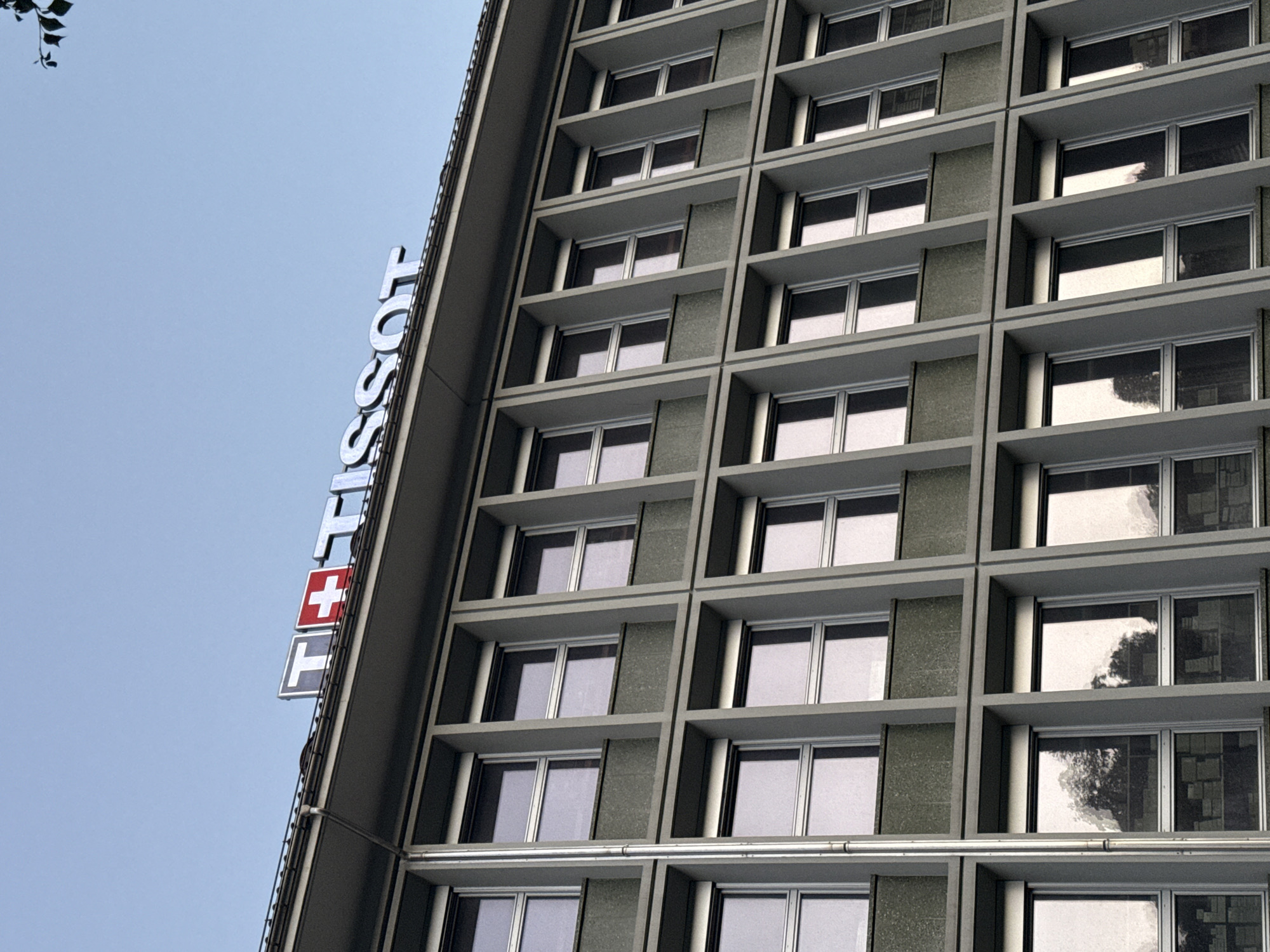 The image shows the Tissot headquarters building in Le Locle, Switzerland. The building is a multi-story structure with a facade composed of repetitive rectangular windows set within a grey grid. Above the windows, the Tissot logo is visible, featuring a Swiss flag symbol and the word "TISSOT" in illuminated letters. The sky is a clear, pale blue. There are no people visible in the image. The overall impression is one of a modern, functional corporate building under a bright sky.