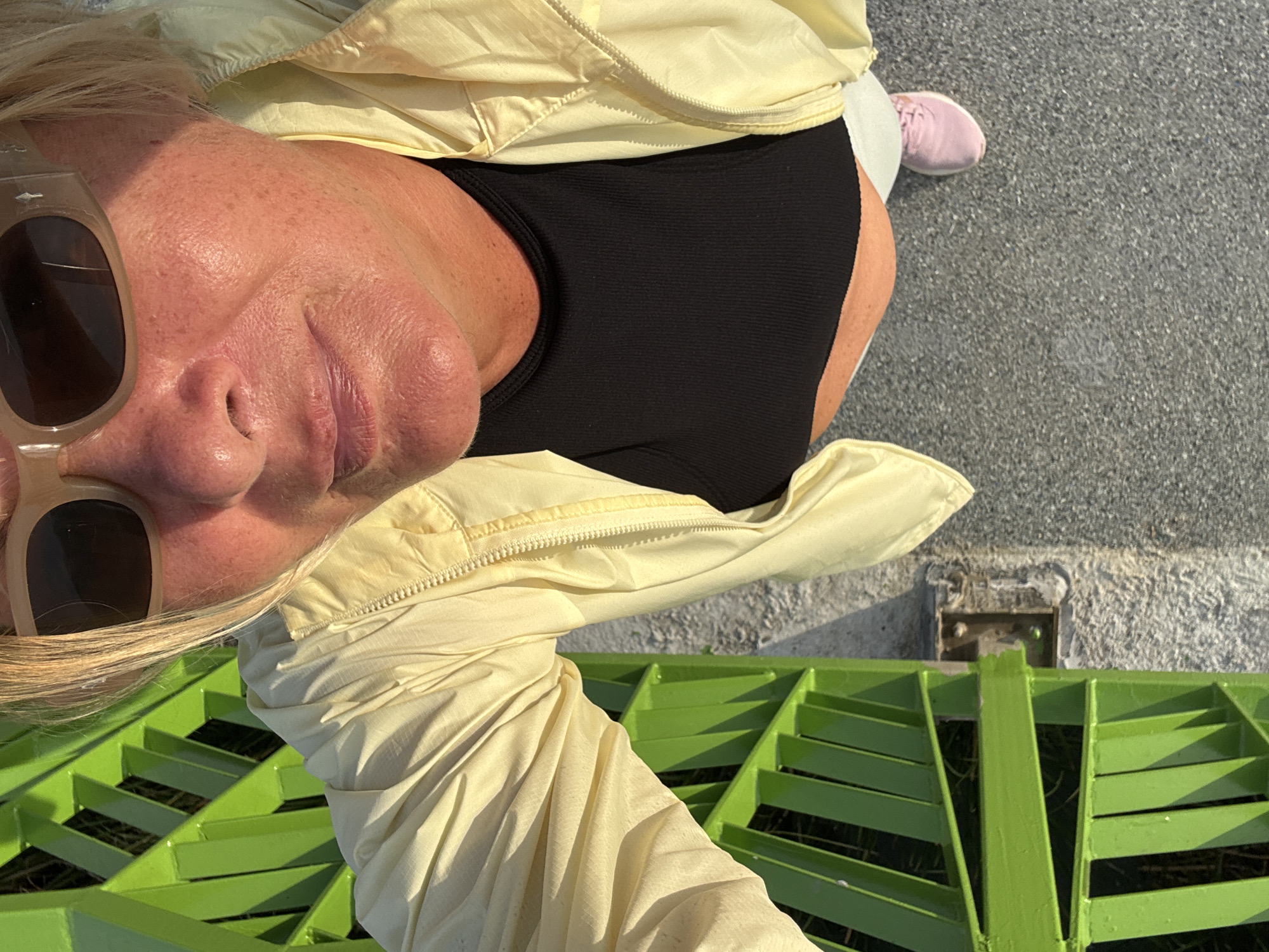 A high-angle, rotated view shows a woman in บ้านบางรักษ์, Thailand. She wears tan-tinted sunglasses, a pale yellow jacket, and a black top. Parts of a lime green, slatted structure are visible near her. A pink shoe is seen. The surrounding area appears to be concrete.