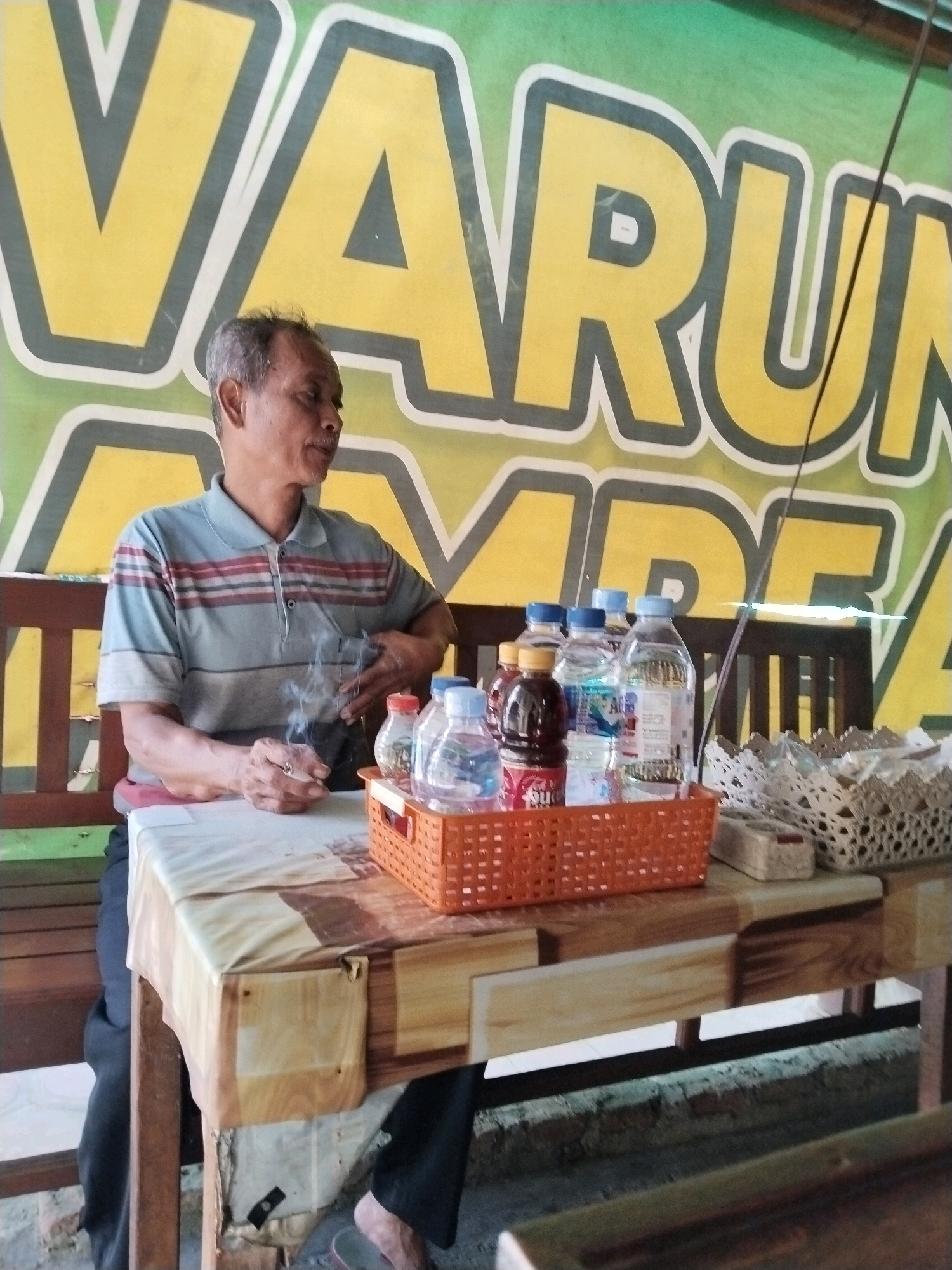 A middle-aged man with graying hair and a mustache is seated at a table, smoking a cigarette. He wears a short-sleeved collared shirt with horizontal stripes in gray, red, and black. The table is covered with a plastic tablecloth and has an orange woven basket holding several bottles of drinks, including water and what appears to be a dark-colored beverage. To the right of the basket are two intricately woven white trays or containers. The man's left hand rests on the table, and his right arm is bent as he holds the cigarette, with smoke rising from it. In the background, a large sign with yellow lettering on a green background is visible, featuring large, stylized letters that appear to spell "VARUNG...". The setting suggests a casual outdoor or semi-outdoor establishment, potentially a small eatery or convenience store in Trenggalek, Indonesia, given the sign's likely local language or branding. The table itself is made of wood, with some exposed brickwork visible below it.