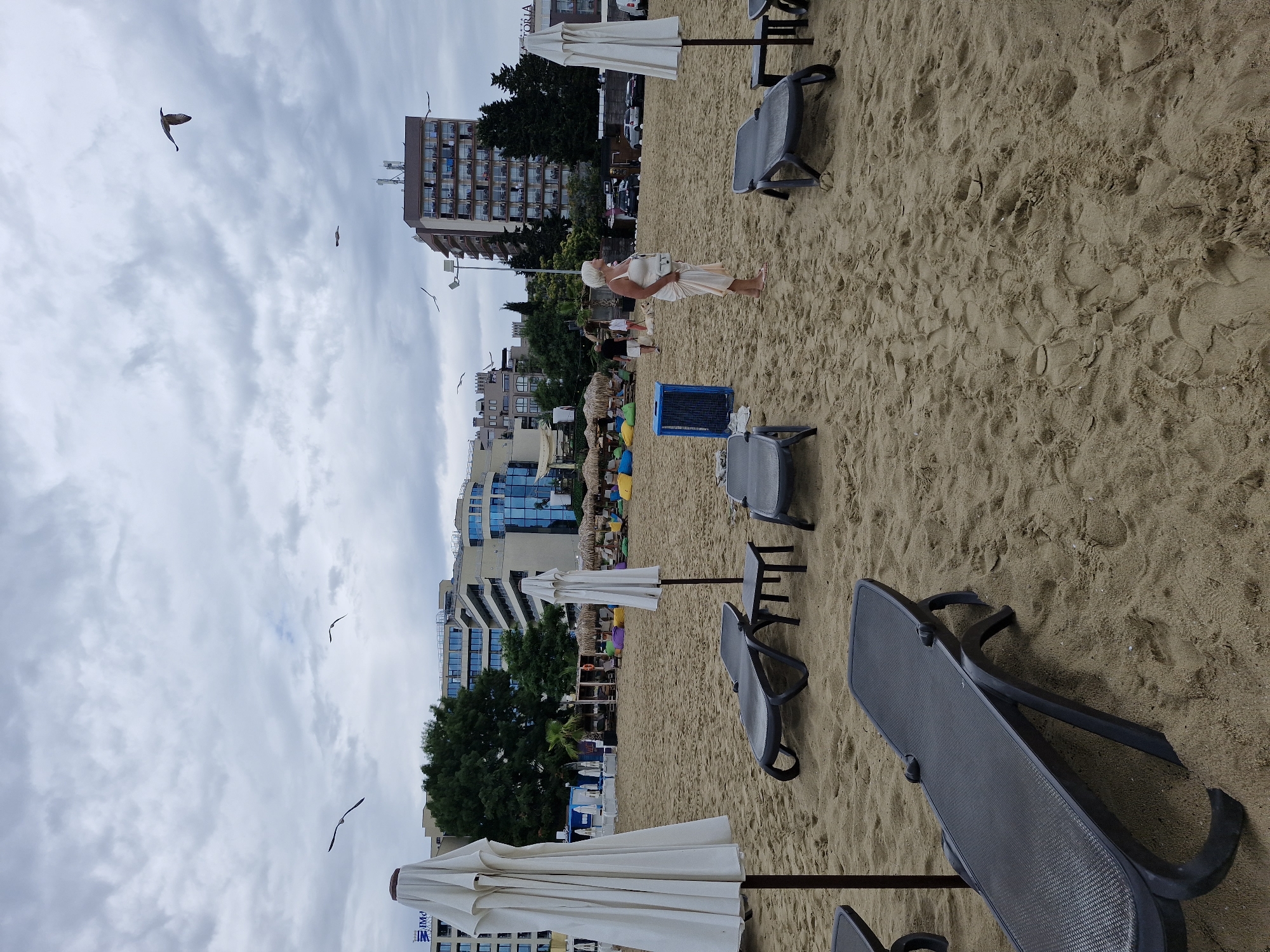 The image depicts a beach scene in Nesebar, Bulgaria, under a cloudy sky. In the foreground, several empty black plastic lounge chairs and white beach umbrellas are set up on the sand. A woman with short blonde hair, wearing a light-colored dress and white purse, stands in the middle ground, facing right and looking away from the camera. Behind her, a blue garbage bin sits near other beach furniture, including colorful beanbag chairs and thatched-roof umbrellas. Further back, various multi-story buildings, some with balconies and glass windows, line the coastline. Several seagulls are visible in the sky, with one in flight towards the upper right corner and another to the left. The sand is textured with footprints and the general atmosphere suggests a partly cloudy day at a resort.