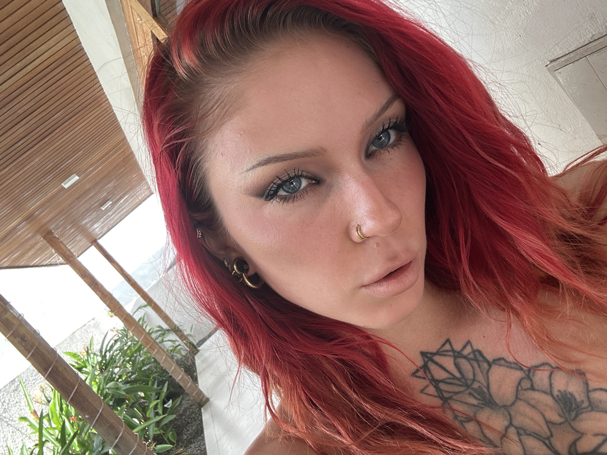 A close-up, low-angle, portrait-style photograph shows a person with vibrant red hair and striking blue eyes. The individual's head is tilted, with their gaze directed towards the viewer. They are wearing subtle makeup, including eyeliner and mascara, and have a nose ring. A portion of a floral tattoo is visible on their chest and shoulder. The background suggests an outdoor setting, possibly a covered patio area, with elements of tropical foliage and a wooden-slatted ceiling. The lighting is bright, indicating daytime. The overall composition is intimate, focusing on the subject's face and expression.