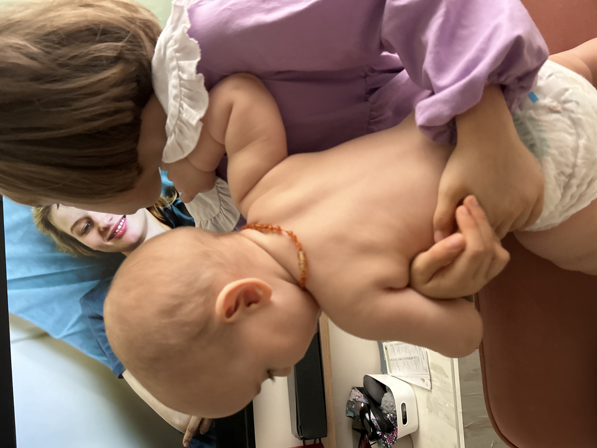 An indoor, sideways-oriented photograph captures a tender interaction between two children, observed by an adult. A young child, with light brown hair, wearing a long-sleeved lavender dress with white ruffled trim, is holding a baby. The baby is mostly bare-chested, wearing only a white diaper and an amber beaded necklace around its neck, and is positioned almost upside down relative to the viewer, being held by the older child's hands around its waist and bottom.
In the background, a smiling adult woman with blonde hair is partially visible, lying or reclining on a surface covered with blue fabric, possibly a medical exam table or a bed. Her smile suggests warmth and contentment as she looks towards the children.
Notable objects in the scene include a black speaker on a stand, a white plastic container filled with assorted items, and some papers or documents affixed to a light-colored wall or partition. Part of a brown, upholstered surface, possibly a chair or table, is also visible. The lighting suggests an indoor setting during daytime, with no specific time of day or weather discernible. No visible text is clearly readable in the image or its crops. While the location context is given as Moscow, Russia, there are no specific visual cues within the image itself to identify this city.