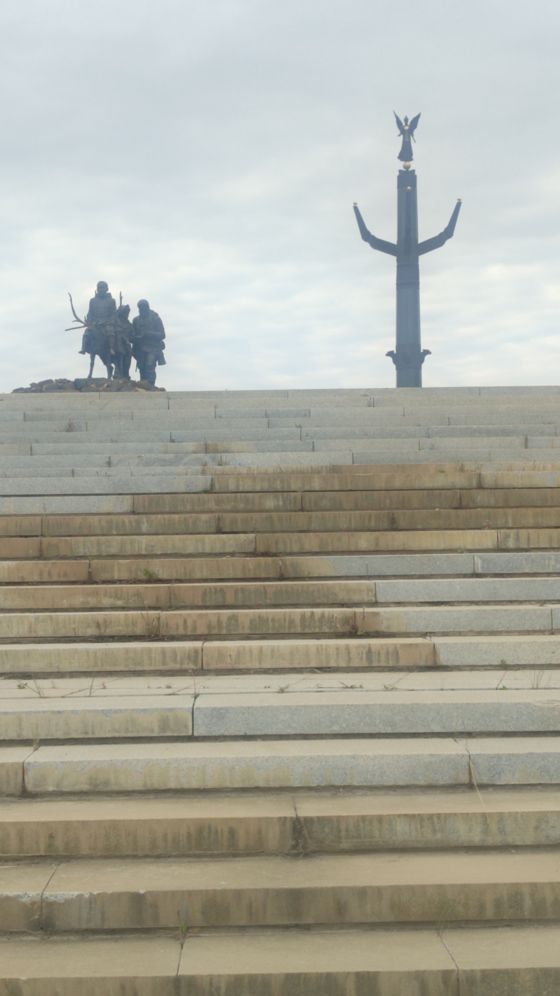 The image shows a view looking up a wide, outdoor stone staircase towards a monument. The monument consists of a tall, dark, columnar structure topped with a statue of an winged figure. To the left of the main monument, on the upper level of the staircase, there is a bronze equestrian statue of three figures, one on horseback and two standing. The sky is overcast. The location is identified as Mirny, Russia, and the monument is likely the "Miracle of Mirny" monument, also known as the "Angel of Mirny," dedicated to the diamond miners of Yakutia. The staircase is made of light-colored stone blocks, some showing wear and small plants growing in the cracks. The overall scene is a public monument viewed from below.