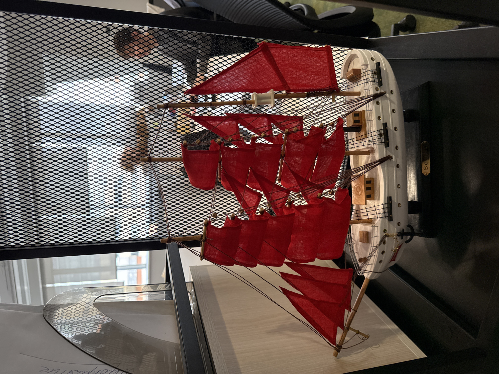 The image depicts a model ship with red sails displayed on a shelf inside a black metal grid structure, possibly a cabinet or storage unit. The ship model features a white hull and multiple red cloth sails, along with wooden masts and rigging. The shelf the ship sits on is a light-colored, possibly wooden, surface. The surroundings suggest an indoor environment, potentially a home or office setting. Through the grid of the storage unit, a window or source of daylight is visible. Other objects, including shoes and documents, are present in the background. The location in the city is unknown.