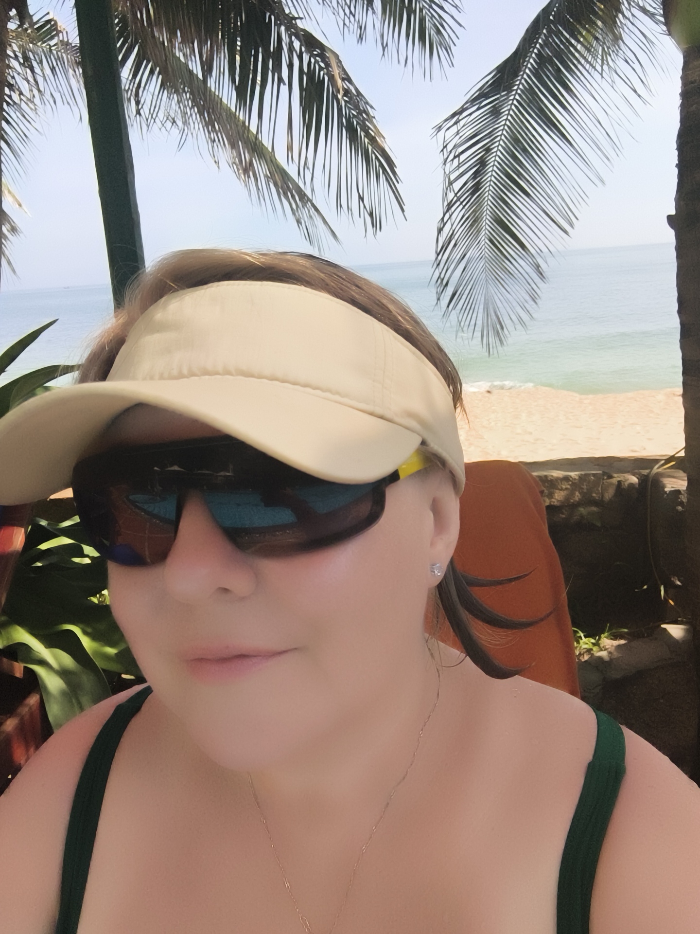 A close-up, selfie-style photograph captures a woman in a tropical setting. She is wearing a light-colored visor and large, dark sunglasses that reflect the sky and palm trees. Her hair, a medium brown, is pulled back and partially visible beneath the visor, with a few strands escaping and framing her face. She is also wearing a dark green swimsuit strap visible on her shoulder and a delicate necklace.
The background depicts a serene beach scene. Lush green palm fronds frame the top and right side of the image, with their shadows falling across the scene. Beyond the palm trees, a calm, light blue ocean meets a sandy beach. The sky is a clear, bright blue, suggesting a sunny day. The overall atmosphere is relaxed and suggests a vacation or leisure activity.
The woman's face is angled slightly towards the camera, and her lips are softly closed, giving a relaxed and pleasant expression. The lighting suggests it is daytime, likely mid-morning or afternoon, given the brightness of the sun and the shadows cast. The presence of palm trees and the ocean strongly indicate a tropical beach location. There is no visible text in the image.