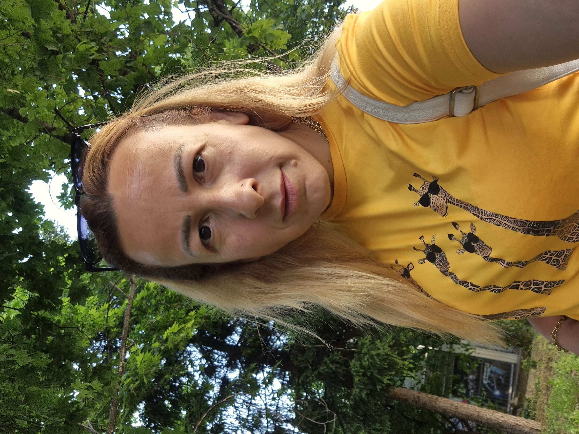 The image presents a close-up, tilted portrait of a woman outdoors during the daytime. She has long, blonde hair, dark eyes, and a neutral to slightly contemplative expression, looking directly at the camera. She is wearing a bright yellow t-shirt adorned with stylized black and white giraffe patterns. A white strap, possibly from a bag or garment, crosses over her right shoulder (viewer's left), and a delicate gold chain is visible around her neck. Sunglasses are perched on top of her head.
The background is dominated by dense green foliage and tree branches, indicating a natural outdoor setting like a park or garden. Bright, natural light illuminates the scene, suggesting clear or partly cloudy weather. In the distance, through the trees, a faint structure resembling a building with a window can be vaguely discerned. The giraffe patterns on her shirt feature intricate, decorative elements that resemble tribal art or illegible text within their design. The overall atmosphere is casual and relaxed.