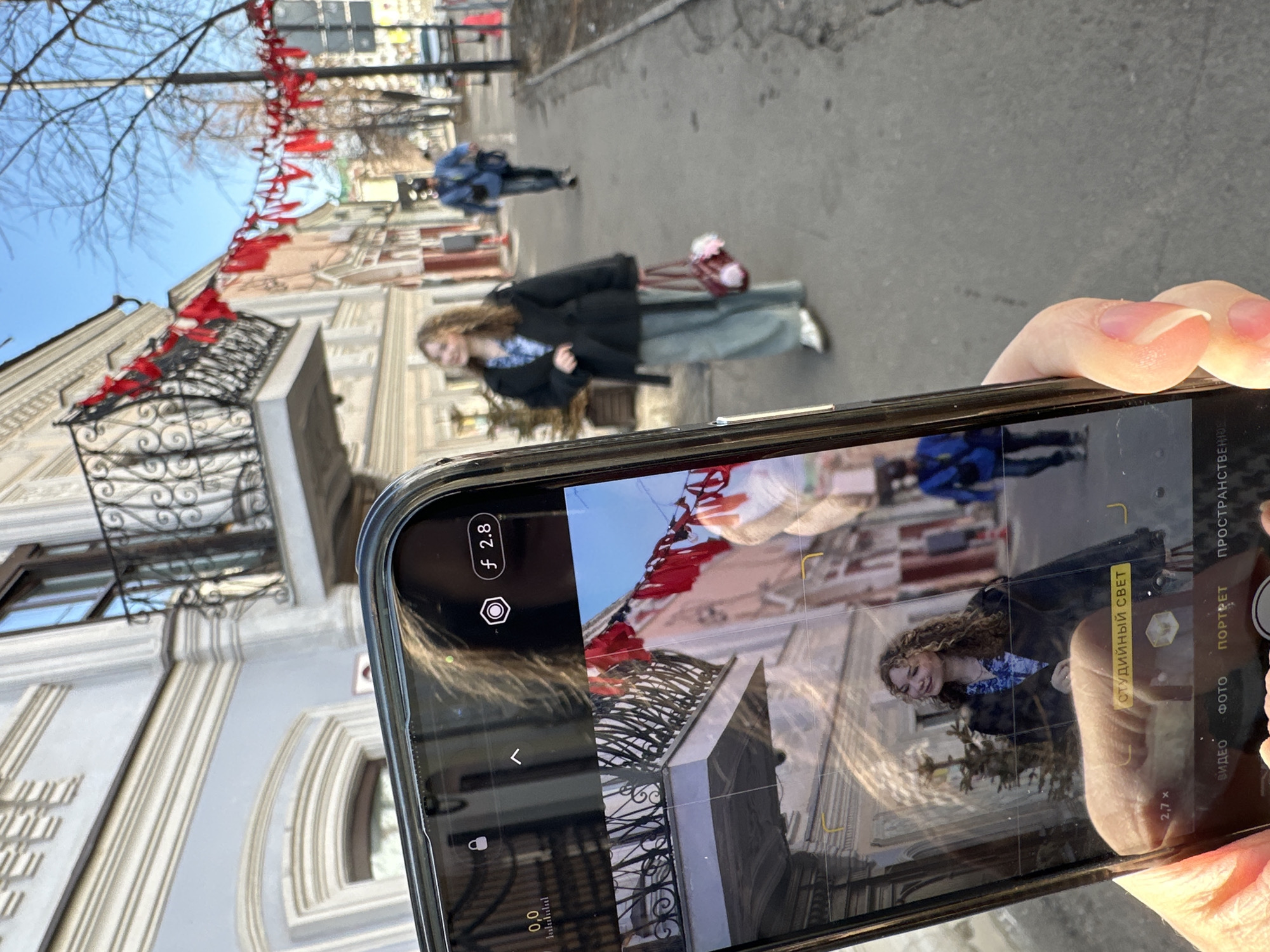 A right hand, manicured with pink polish, holds a black-cased smartphone in the bottom-left portion of the image. The phone's screen displays a live camera feed in portrait orientation, showing a young woman with curly light brown hair, smiling towards the right, wearing a dark jacket, a blue patterned shirt, and holding a red handbag. The camera interface displays "f 2.8" and the Russian text "СТУДИЙНЫЙ СВЕТ" (Studio Light).
The scene beyond the smartphone is a sunny daytime view of Bauman Street, a pedestrian thoroughfare in Kazan, Russia. A young woman, matching the one on the phone screen, walks left on the paved sidewalk. She wears a dark overcoat, a blue and white patterned shirt, light blue wide-leg pants, and white sneakers, carrying a burgundy handbag with two fluffy pom-poms on her left arm. Her gaze is directed slightly upwards and to her right. Further down the sidewalk, another individual walks away from the viewer, dressed in a blue jacket, dark pants, and a dark cap.
The street is lined with multi-story historic buildings. The building on the immediate left is light grey, featuring white ornate cornices, window frames, and a prominent black wrought-iron balcony. Other buildings further down the street exhibit various pastel colors. Red fabric streamers are strung across the street between buildings. Bare trees are visible along the sidewalk, and the sky is clear blue.