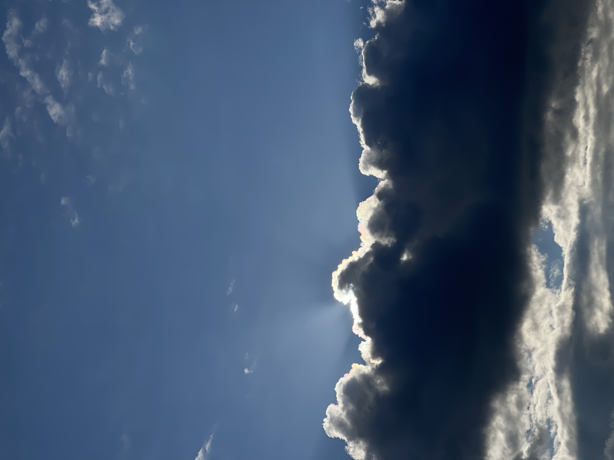 The image displays a dramatic sky scene with a clear blue expanse at the top, gradually transitioning to a dense, dark cloud formation that dominates the lower portion of the frame. Sunlight pierces through a gap in the clouds, creating visible sunbeams that illuminate the edges of the clouds with a bright, iridescent glow, hinting at a prismatic effect. The clouds themselves are varied, with the upper edge of the dark formation appearing bright and textured where the sun hits it, while the bulk of the cloud is a deep, ominous dark blue-gray. The lower part of the image shows more scattered, lighter clouds, some catching the sunlight and appearing bright white, while others are shadowed. The overall impression is one of a dynamic sky, possibly at sunrise or sunset, with a strong contrast between light and shadow. The location is not specified, and no people or specific objects are visible, making the scene a natural landscape focused solely on the sky.