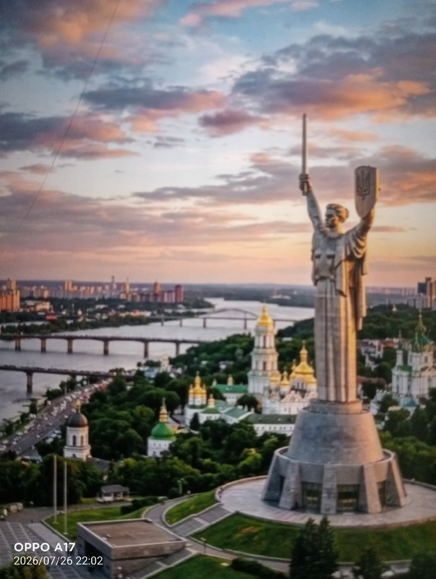 The image shows a panoramic view of a city skyline at sunset. In the foreground, a large statue of a woman holding a sword and shield stands prominently. The statue is made of stone and is mounted on a circular pedestal. Behind the statue, a river can be seen with several bridges crossing it. The city skyline in the distance features numerous buildings of various heights and architectural styles. The sky is filled with clouds, illuminated by the warm hues of the setting sun, casting a golden glow over the scene. The overall impression is one of grandeur and urban development against a dramatic natural backdrop. The image does not contain any recognizable elements that place it specifically in Құмөзек, Kazakhstan.