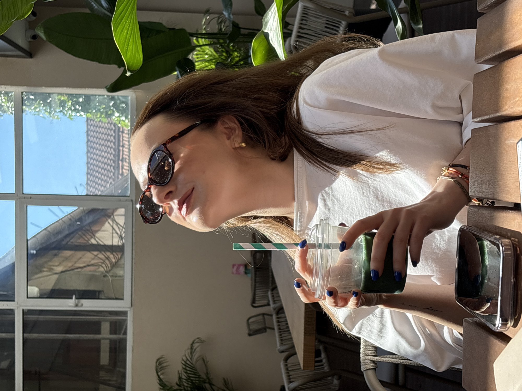 A young woman wearing sunglasses and a white t-shirt holds a clear plastic cup with a green beverage and a striped straw. She is seated at a wooden table, with a smartphone reflecting the green drink in front of her. The background suggests an outdoor cafe setting with tables and chairs visible, and large windows revealing a sunlit sky and buildings. The lighting indicates it is daytime. This scene is at the cafe "City Cafe" in Kragujevac, Serbia.