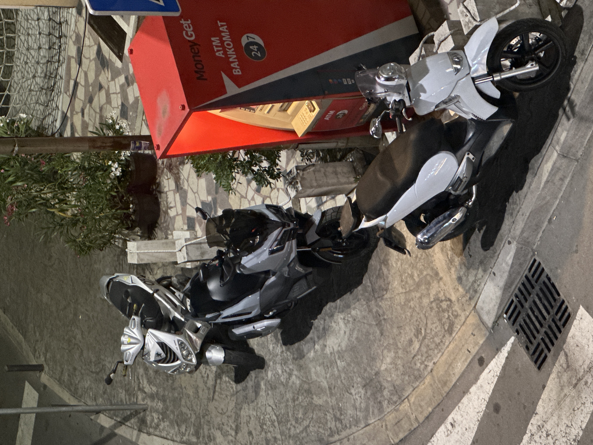 The image captures an outdoor street scene at night, likely in an urban area of Budva, Montenegro. Two scooters are parked on a paved surface next to a vibrant red ATM machine.
On the right, a light-colored scooter, possibly white or light grey, is positioned with its front wheel towards the viewer, showcasing its disc brake and a black wheel rim. Its seat is black. To the left of this scooter, a larger, grey and black maxi-scooter is parked, displaying detailed fairings and a black seat. Both vehicles appear to be in good condition.
Behind the scooters, a prominent red ATM machine stands out. Visible text on its side includes "Money Get," "ATM," "BANKOMAT," and "24/7," indicating its round-the-clock availability. The ATM is brightly illuminated. Behind the ATM and scooters, there's some dense greenery, possibly bushes or potted plants, with small white and pink flowers.
The ground is paved with irregular stone tiles and concrete. On the far right of the image, a section of a street or sidewalk is visible, featuring a dark metal drain grate and white road markings. The overall lighting suggests it is nighttime, with artificial light sources casting distinct shadows from the scooters and other objects onto the pavement. No people are visible in the scene, which appears quiet and still.