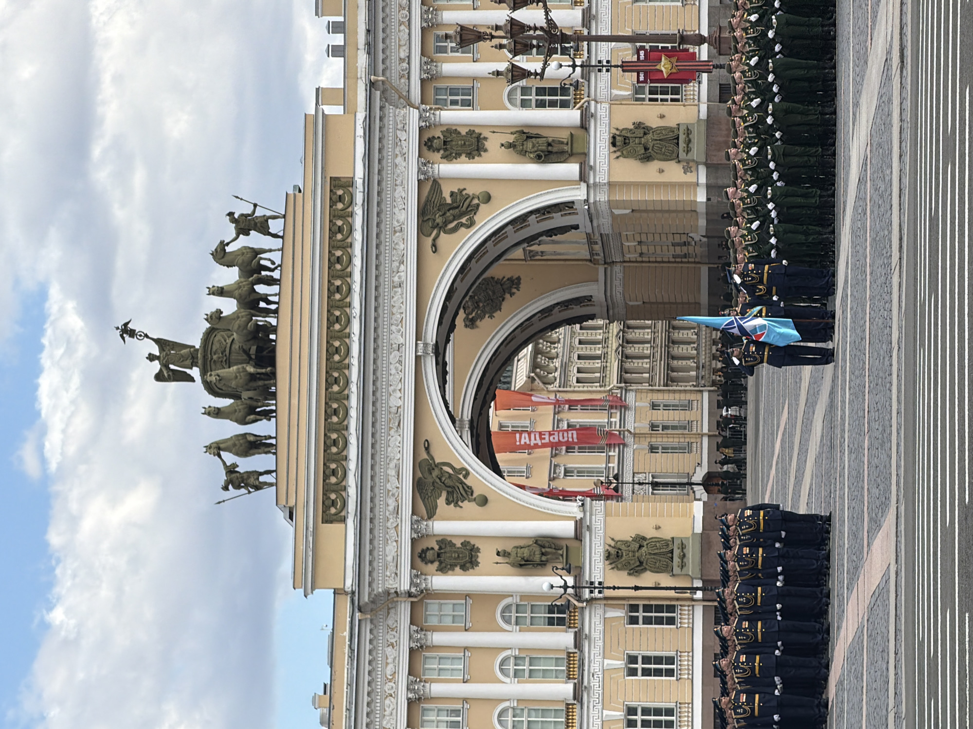 The image depicts a military parade in Saint Petersburg, Russia. In the foreground, uniformed soldiers are lined up, facing right, with a color guard carrying a blue flag towards the front. Behind them is the General Staff Building, specifically the arch topped with a sculpture of a chariot pulled by six horses with two figures. Red banners are hanging from the building. The weather is partly cloudy. The ground is paved with gray stone blocks divided by white lines.