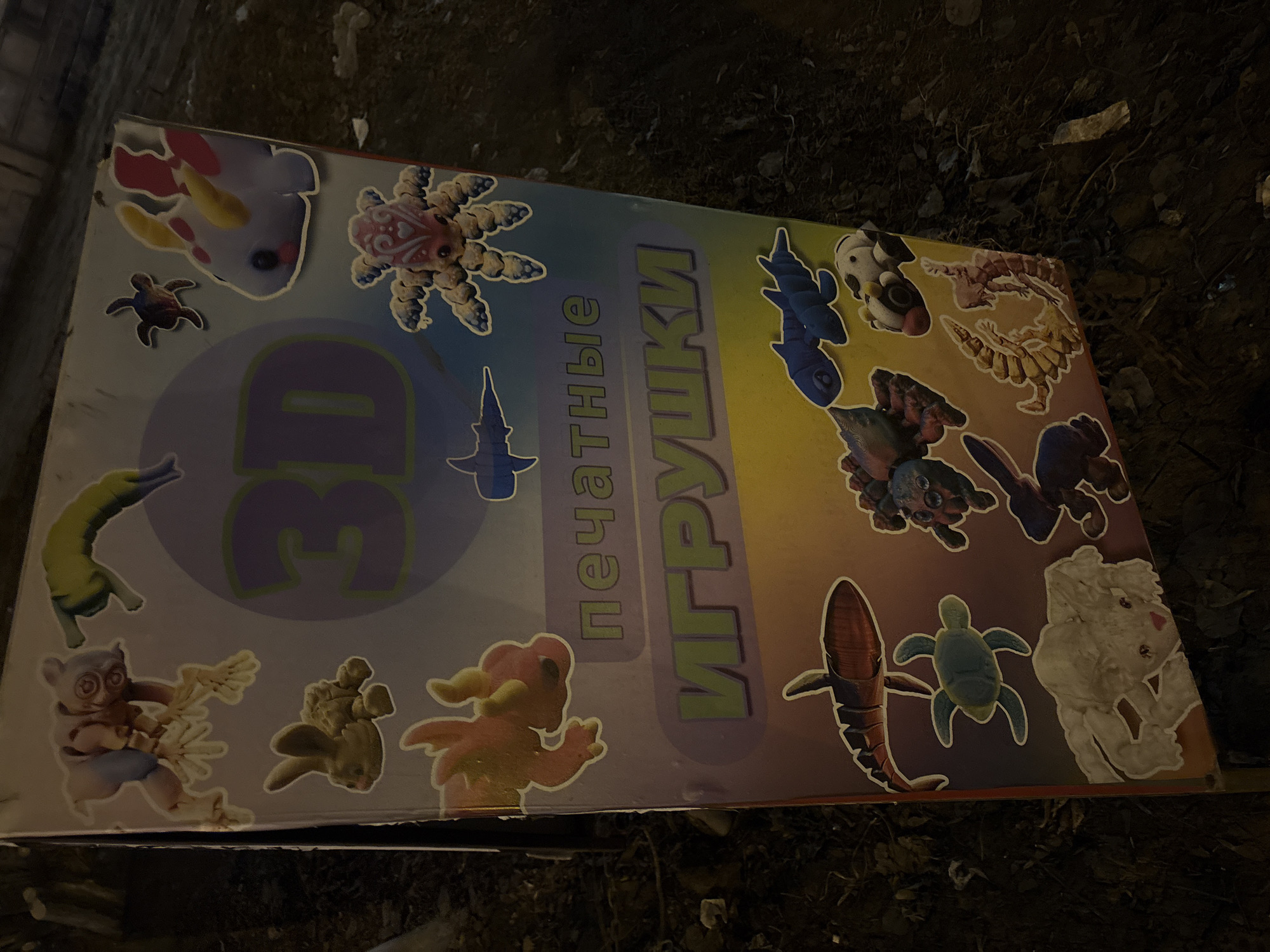 The image shows a promotional sign lying on the ground, likely near a street or sidewalk. The sign advertises "3D printed toys" written in Russian. The sign depicts various 3D printed toy designs, including animals like a shark, turtle, axolotl, spider, rabbit, and other creatures. The environment surrounding the sign consists of dark, unkempt terrain, possibly dirt or debris, suggesting the sign is discarded or has fallen down in a public space. Location cannot be confirmed based on available data.